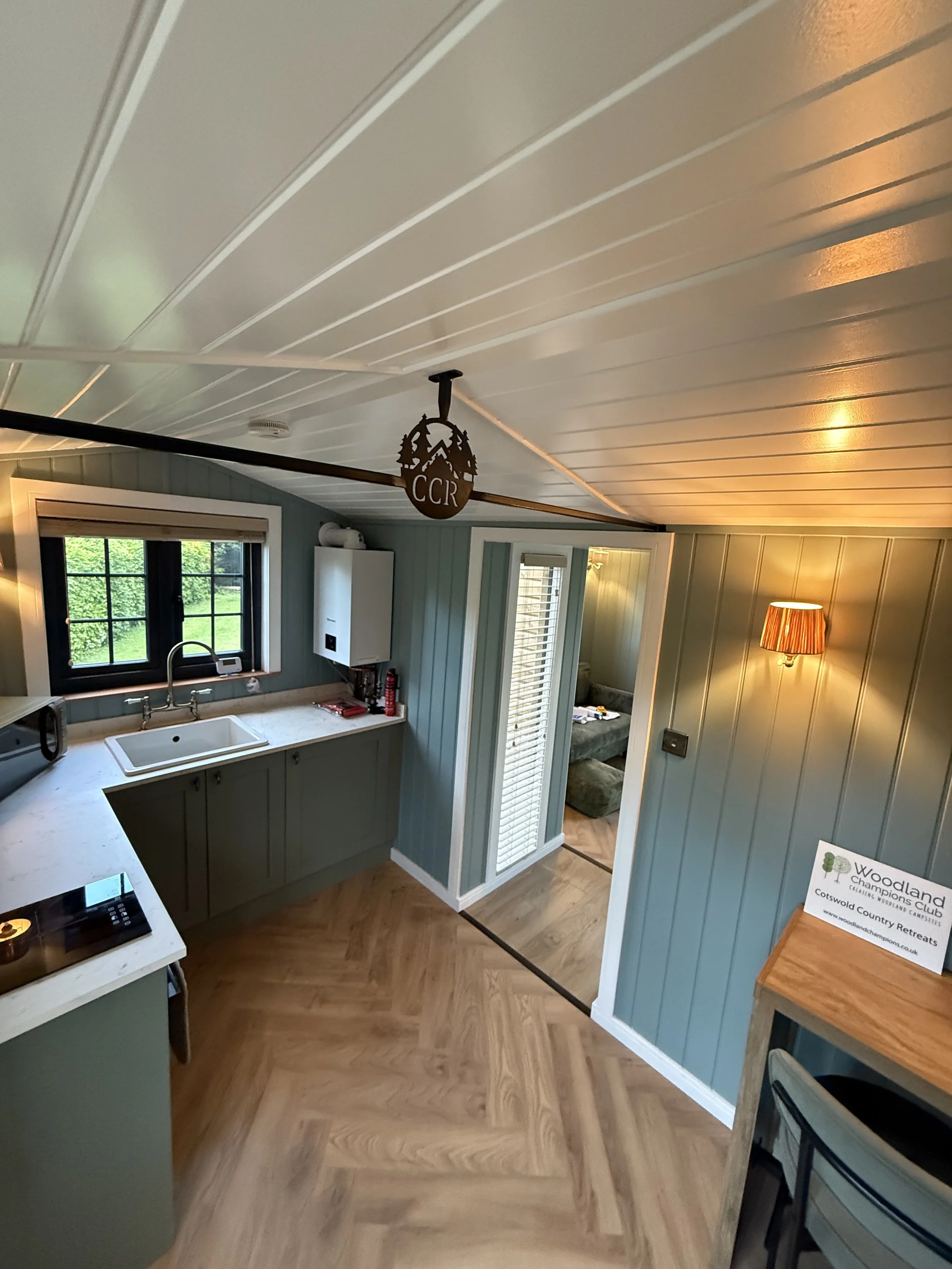 Interior view of a kitchen with a white sink under a window, green cabinetry, and a checkered floor. There is a wooden counter with a lamp and a sign that reads 'Woodland Champions Club'.