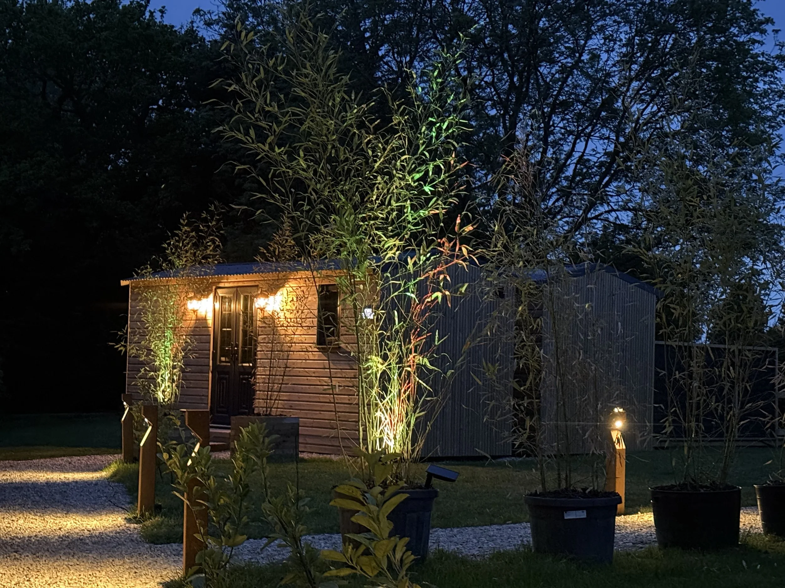 Nighttime scene of a house with exterior lights, surrounded by potted plants and trees, with a dark sky in the background.