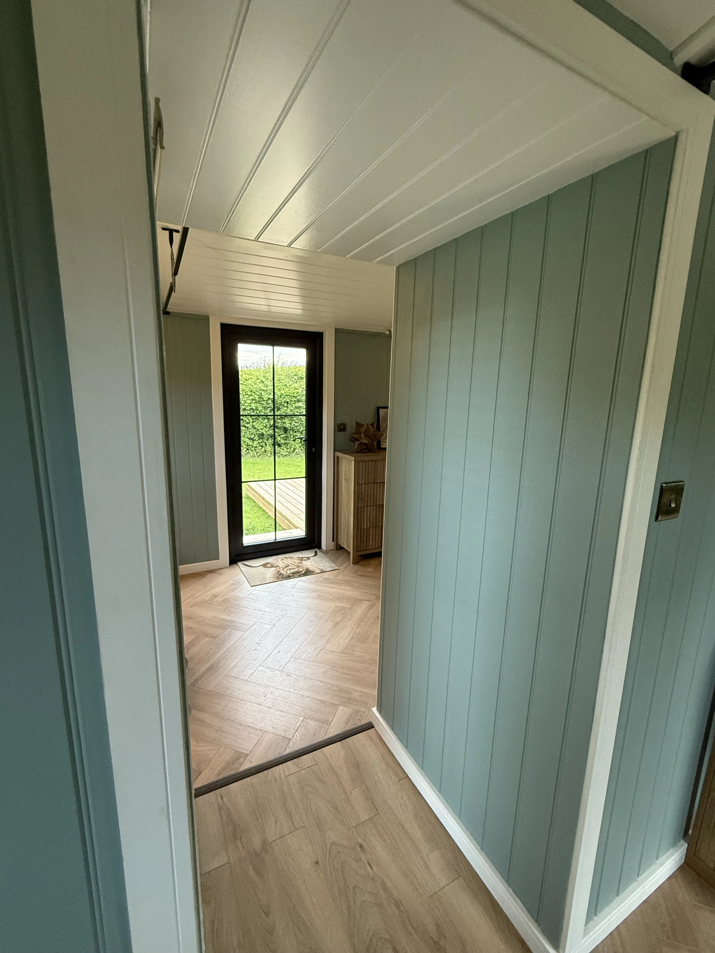 View of a hallway with wood-paneled walls, leading to a door with a large window showing green outdoor scenery, and a small wooden cabinet with decorative items inside a bright, modern home.