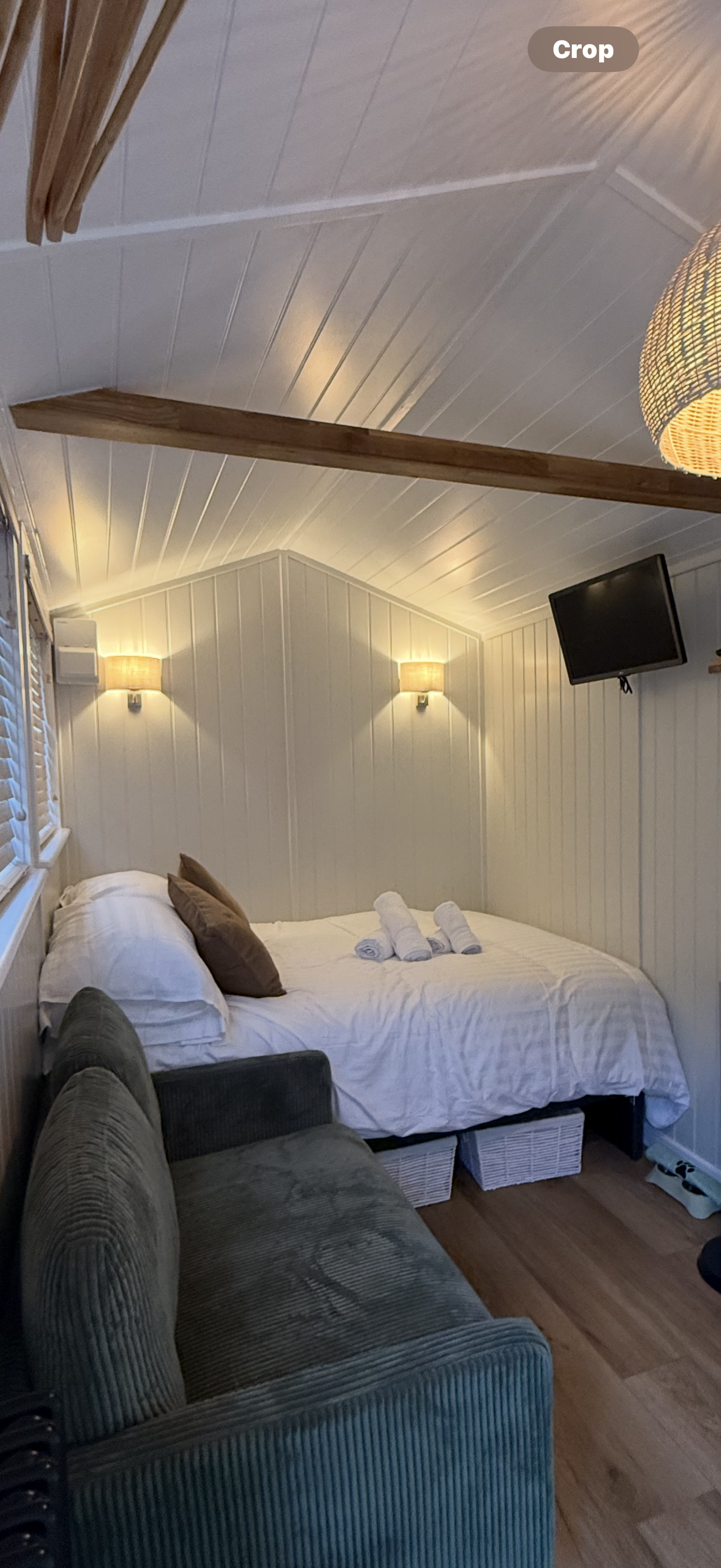 A cozy bedroom with a bed, brown and white pillows, and towels on top. There are wall-mounted lights, a TV, and a window with blinds. A cushioned chair or sofa is in the foreground.