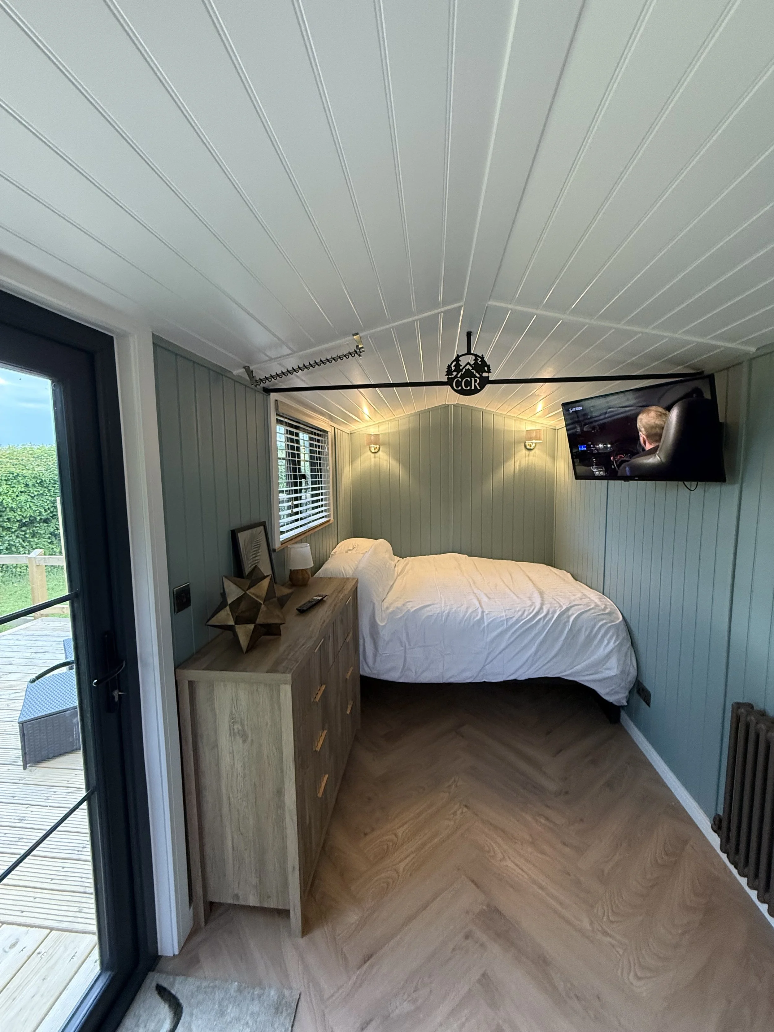 A bedroom with a bed, wooden dresser, wall-mounted TV, and a sliding glass door leading to a deck outside. The ceiling has white paneling and there is soft lighting on the walls.