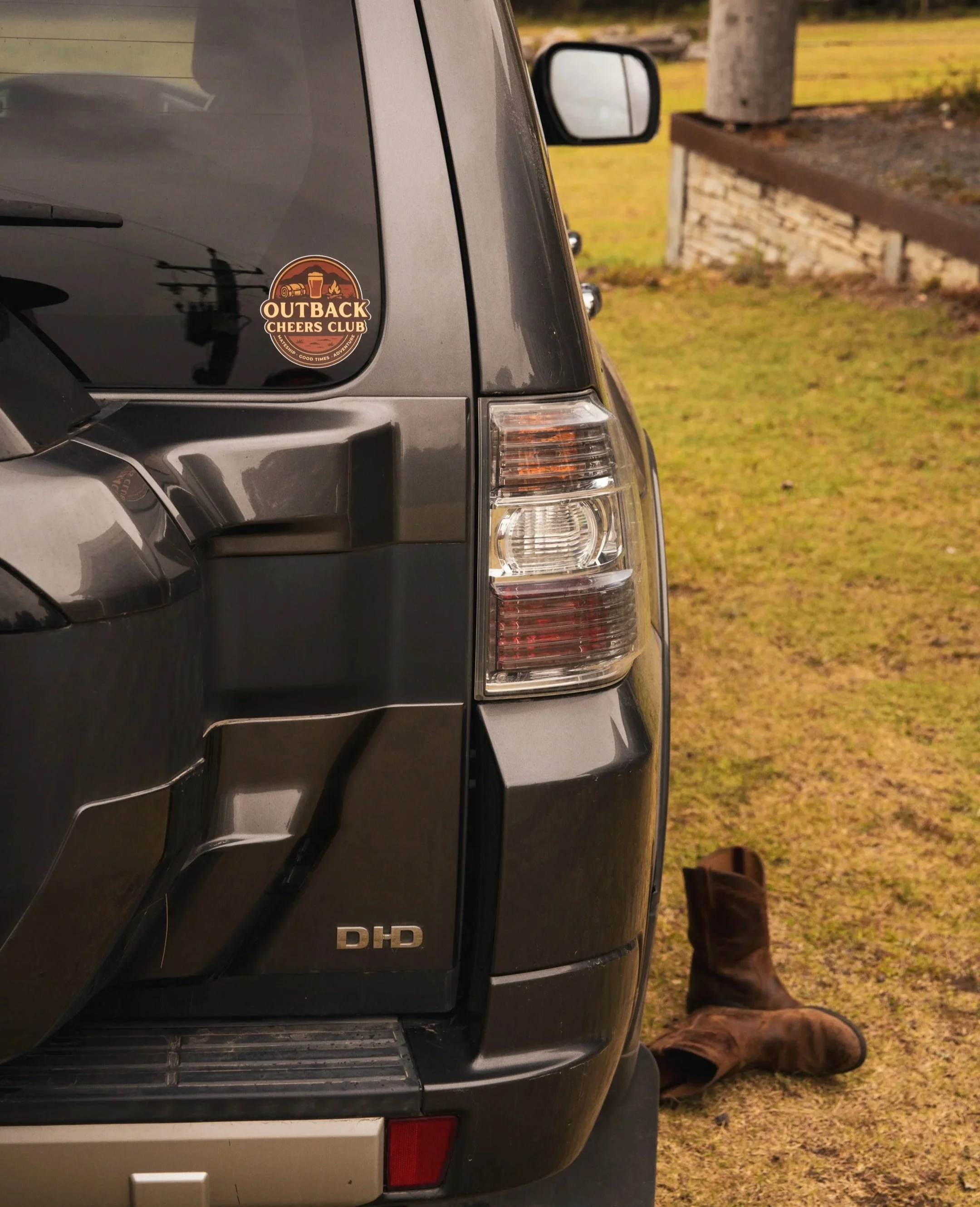 The rear of a grey 4WD with a sticker that says 'Outback Cheers Club' on the back window, a pair of brown cowboy boots on the ground next to the vehicle, and grassy area with a brick border in the background.
