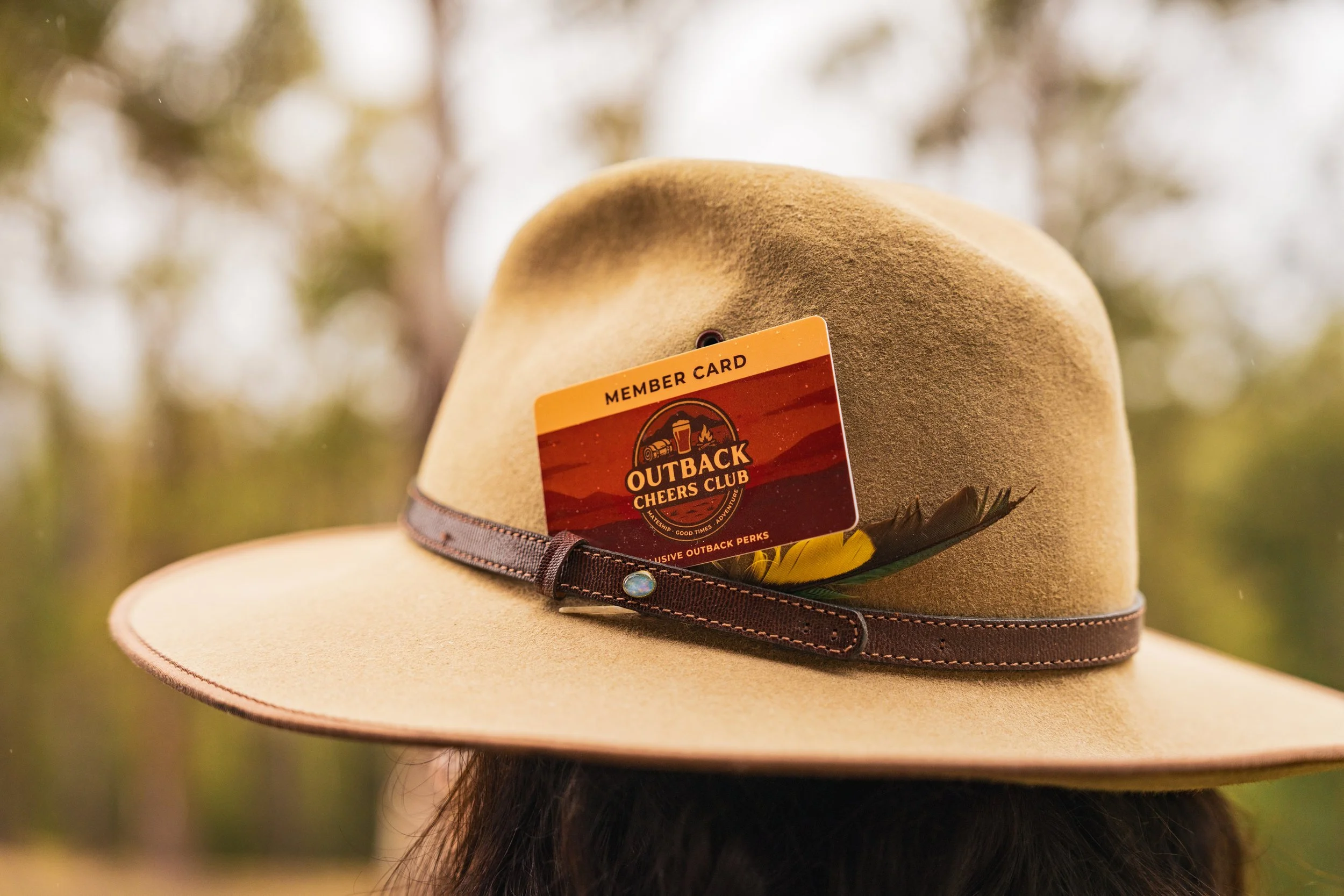 Akubra hat with a brown band and a feather, with an Outback Cheers Club member card tucked into the band, outdoors with blurry trees in the background.