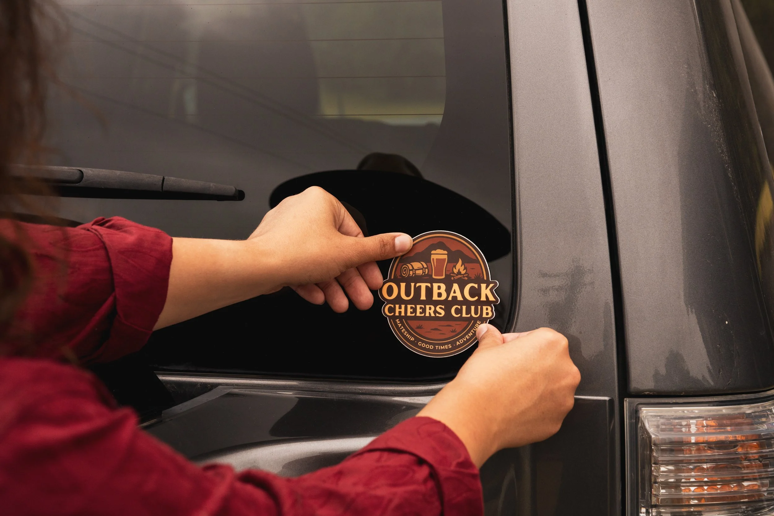 Person applying an Outback Cheers Club sticker to the back of a dark gray vehicle, wearing a red rodeo shirt.