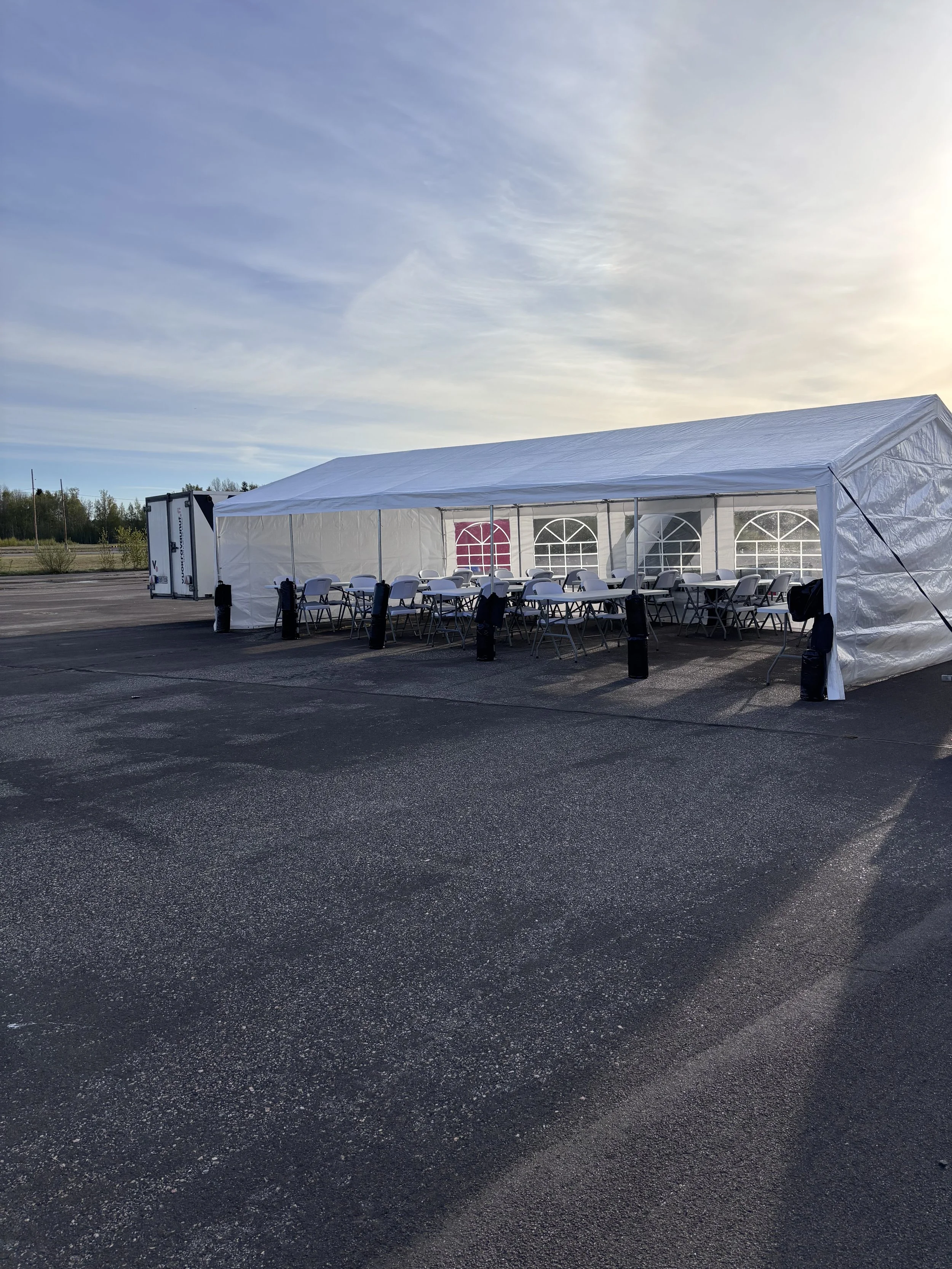 Suuri valkoinen eventtikatto, jossa on valkoiset tuolit ja pöydät, valmistautuu tapahtumaan, paikalla on myös mustia jalkalamppuja, ja taustalla on avoin tilaisuusalue luonnon keskellä aamun tai iltapäivän valossa.