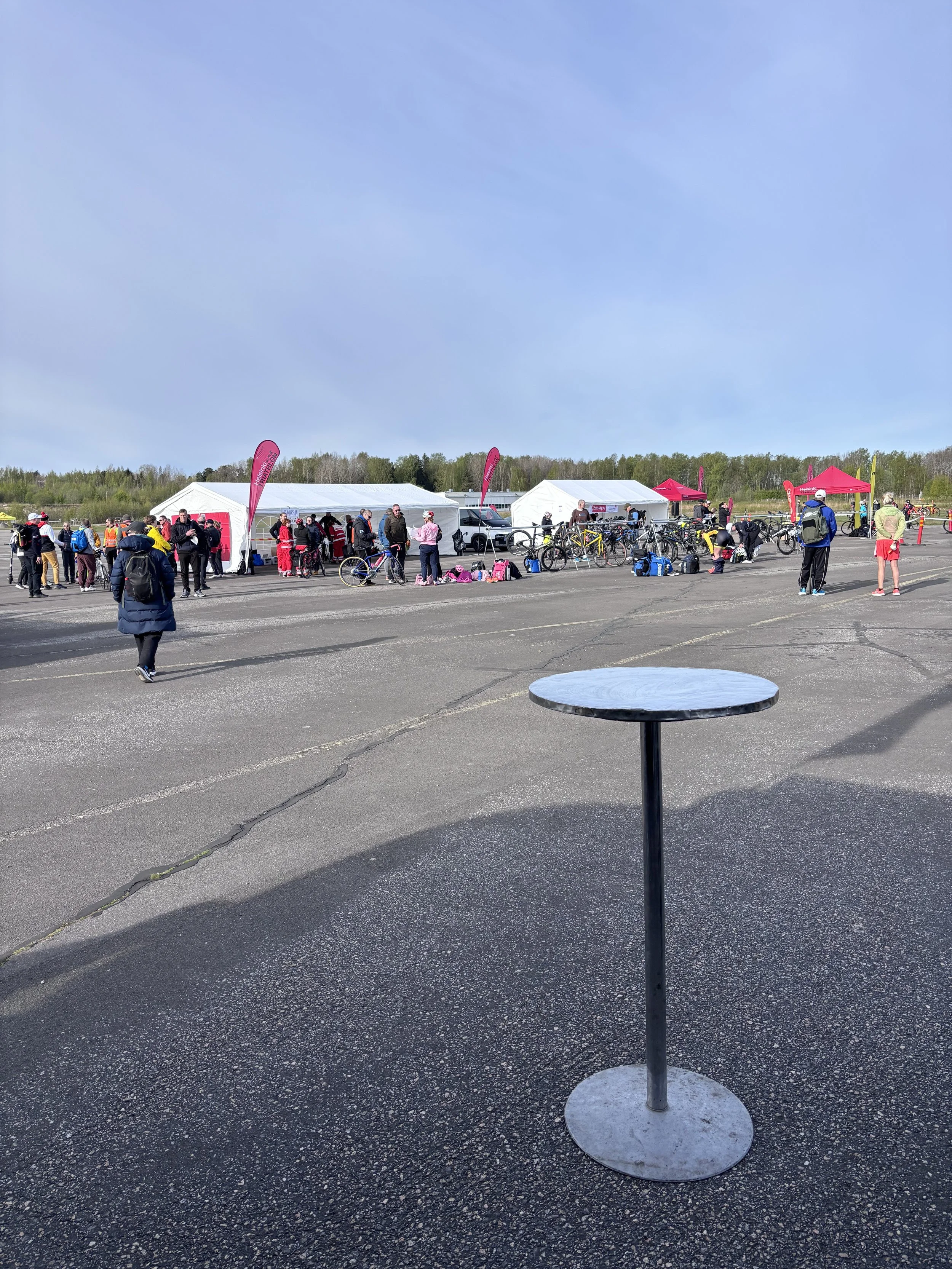 Lehtikokoontumisalueen ulkotapahtuma, jossa on useita ihmisiä ja pyöräparkkeja, telttoja ja jousikankaita, ja taustalla on metsää ja tolppia.