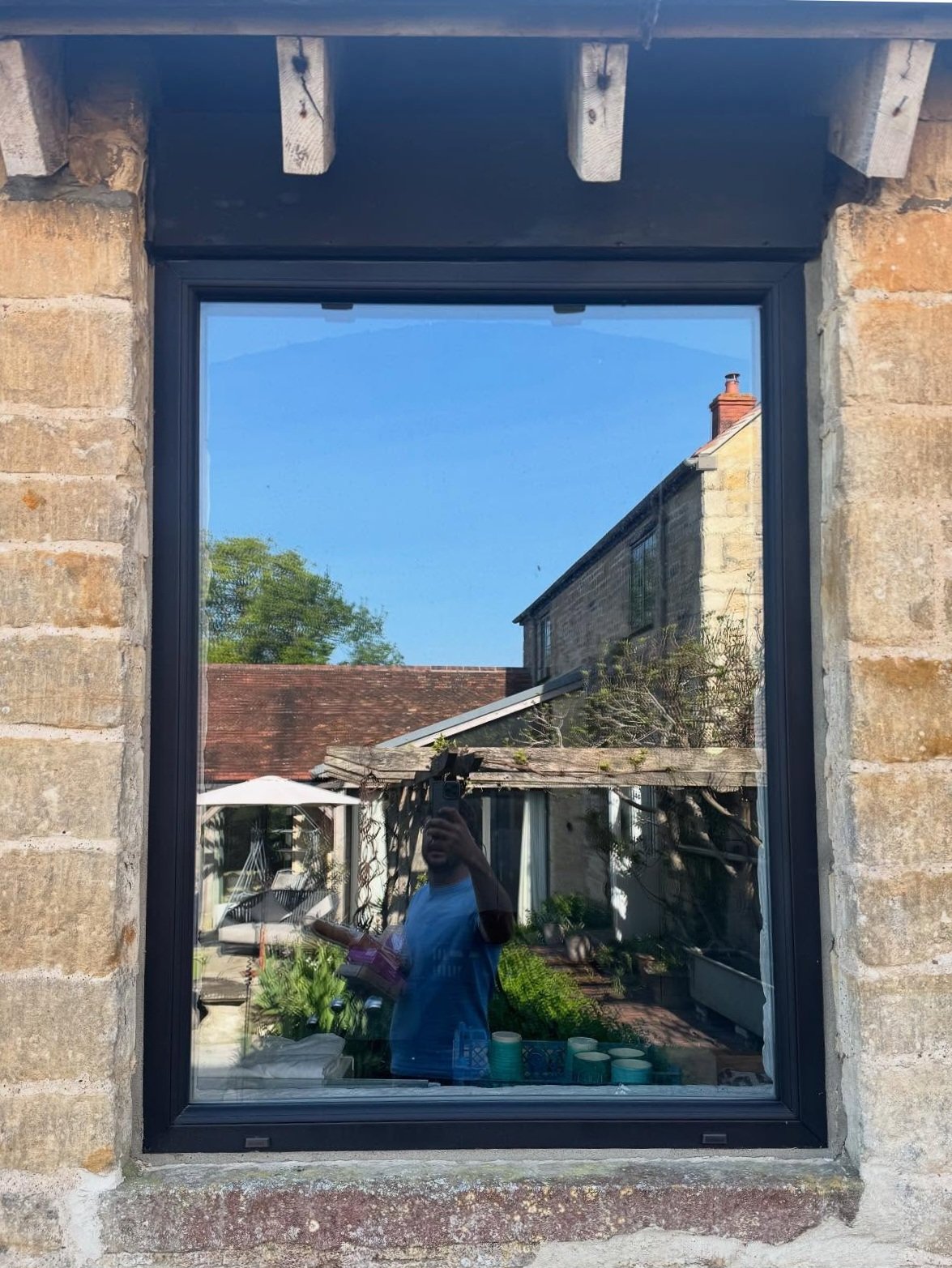 Reflection of a man taking a photo in a window reflecting a garden and neighboring buildings, with blue sky overhead.
