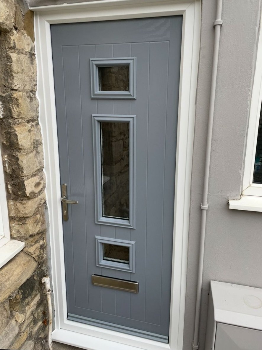 Gray front door with three small rectangular windows and a mail slot, next to a stone wall and a white wall with a window and a pipe.