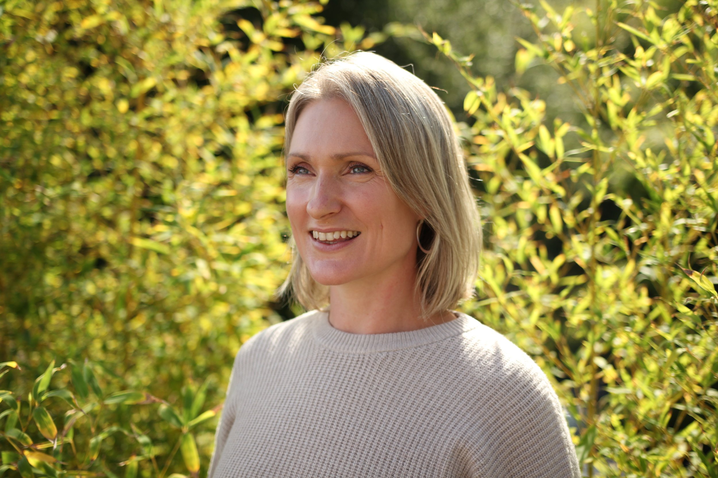 A woman with blonde hair and hoop earrings smiling outdoors with green foliage in the background.