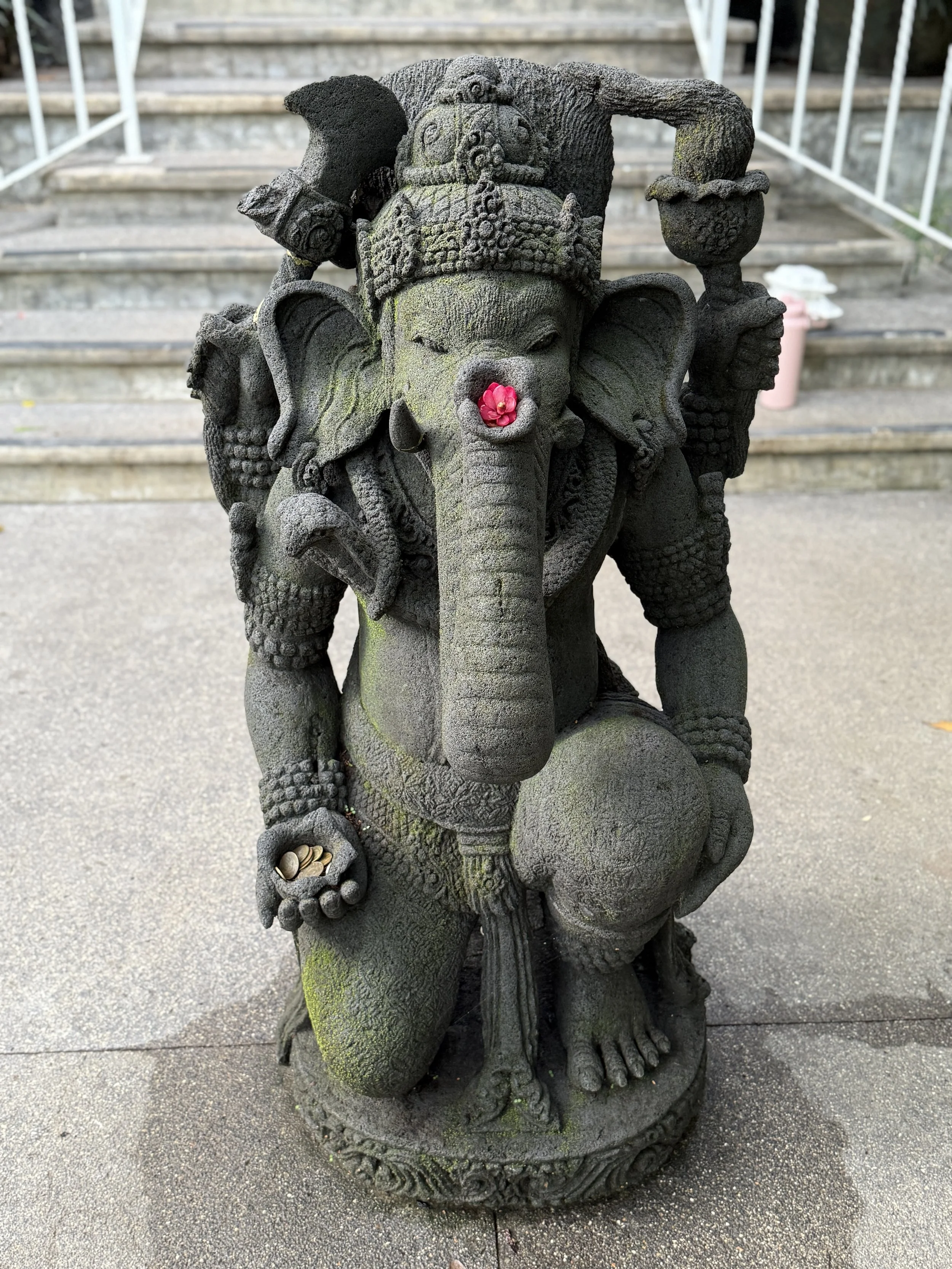 Stone statue of Lord Ganesha, adorned with a pink flower on its nose and coins resting in one hand, set outdoors on a concrete surface.