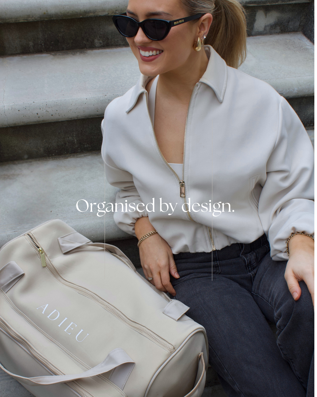 A woman sitting on outdoor steps wearing sunglasses, a white zip-up jacket, and dark jeans. There is a beige bag with the word 'ADIEU' nearby and the text 'Organised by design' overlayed on the image.