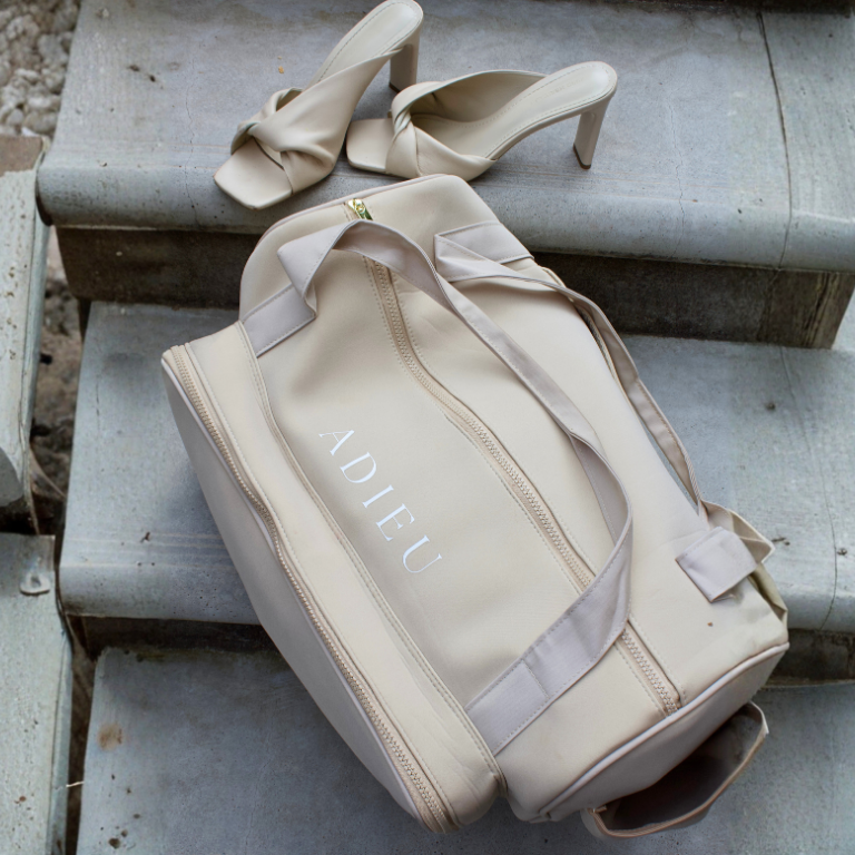 A cream-colored Adieu duffle bag and a pair of cream high-heeled sandals on concrete steps.