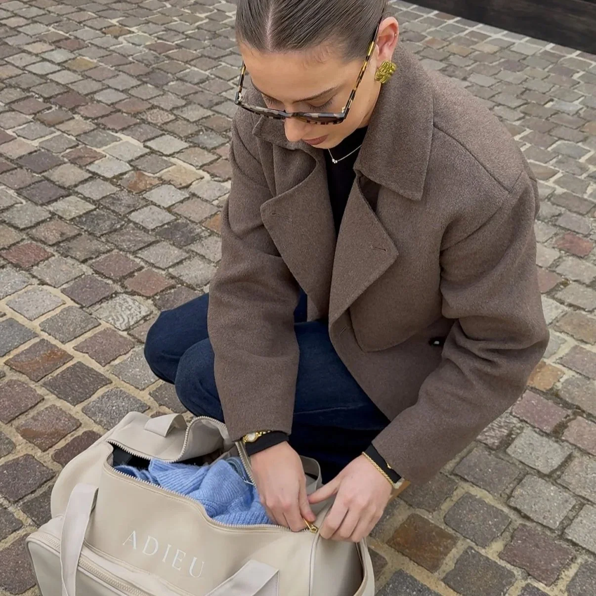 A woman with brown hair, wearing glasses, a brown coat, and dark jeans, is sitting on a cobblestone street packing a beige bag with the word 'ADIEU' on it, containing blue clothing.