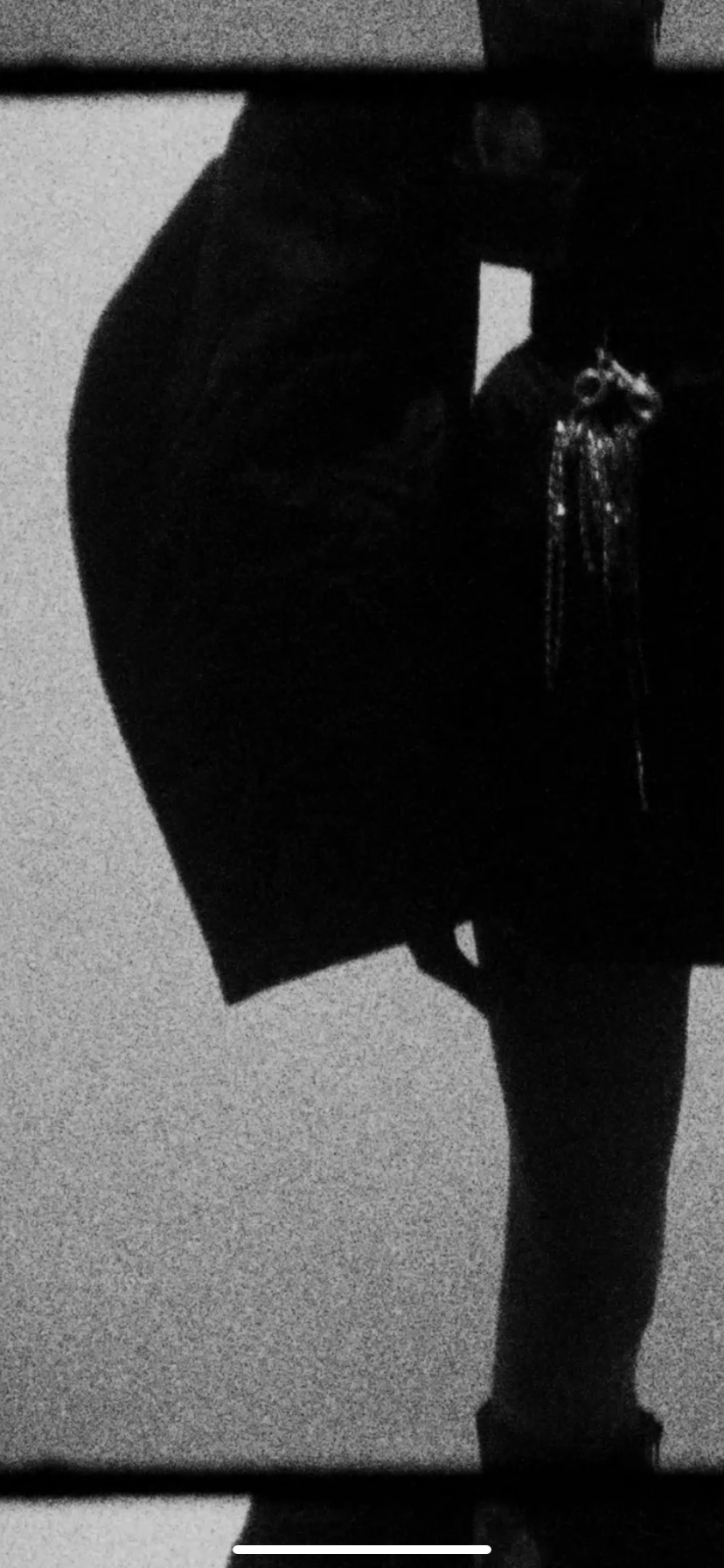 Black and white photograph of a person's lower body, wearing dark clothing and a necklace, standing in front of a plain wall, with a horizontal dark line at the top and bottom of the image.