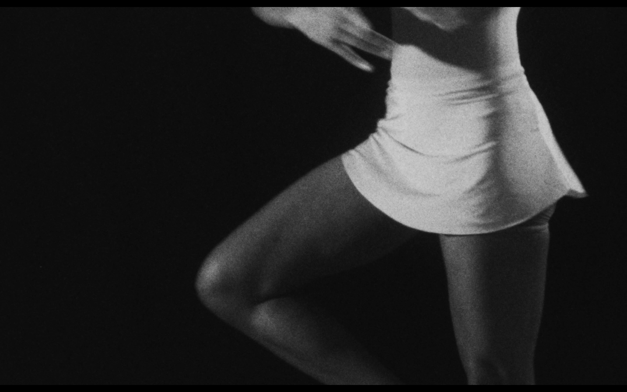 A person kneeling with their head slightly bowed, wearing a light-colored shirt and dark shorts, in a dimly lit environment.