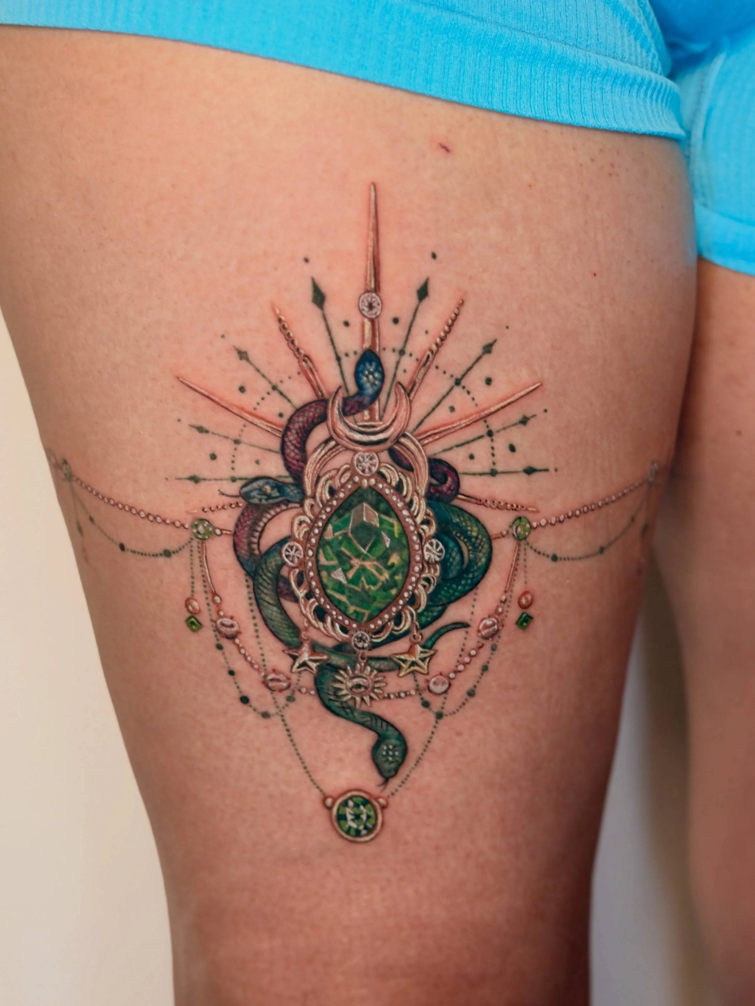 For the woman who wants her tattoo to look like she&rsquo;s wearing the most beautiful piece of jewelry she&rsquo;s never had to take off.

Custom color fine line: emerald gem, crescent moon, snakes, and delicate chain detail across the thigh. Built 