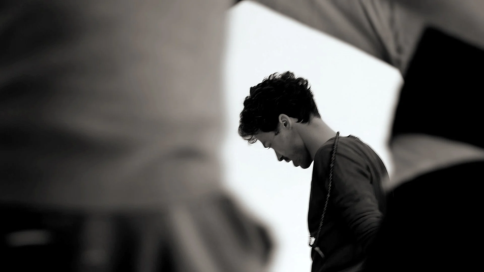 Black and white photo of a young man with short, curly hair looking down, seen from a side profile inside a building or parking garage.