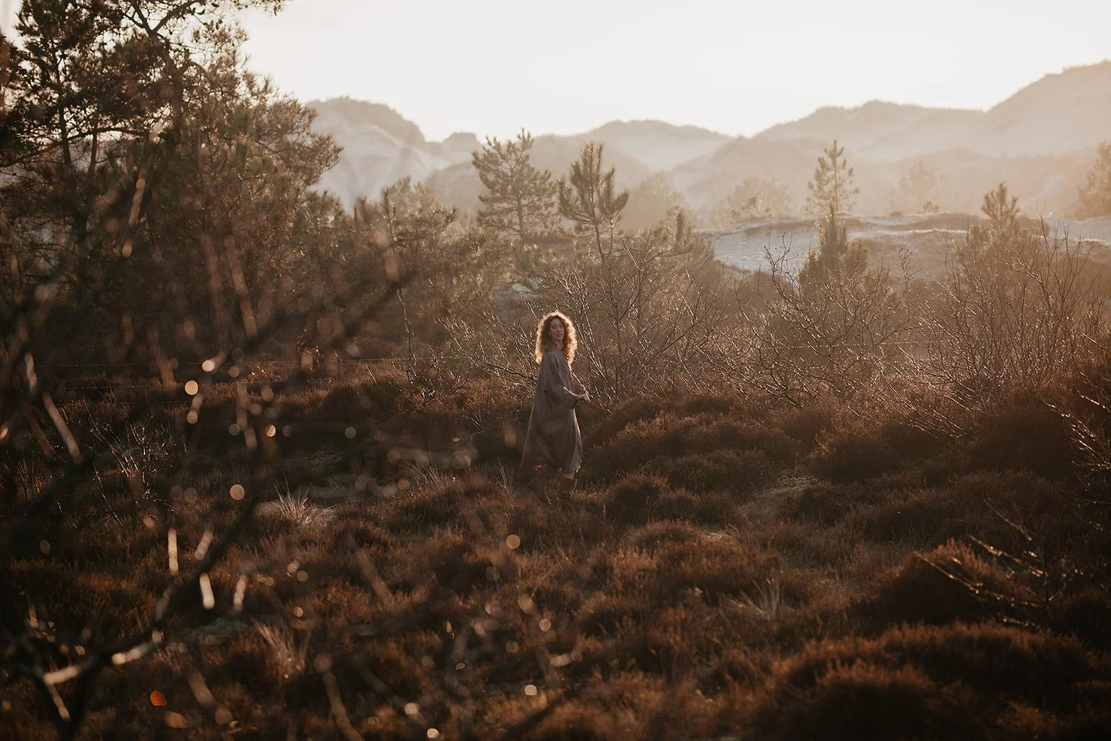 A woman standing in a dry, bushy landscape with mountains in the background, illuminated by warm, golden sunlight.
