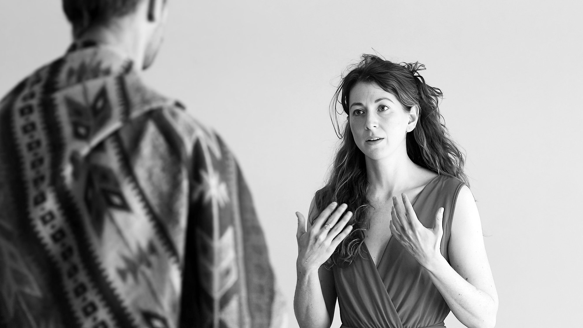 A woman with long hair gesturing with her hands as she speaks to a man in a patterned shirt in an indoor setting.