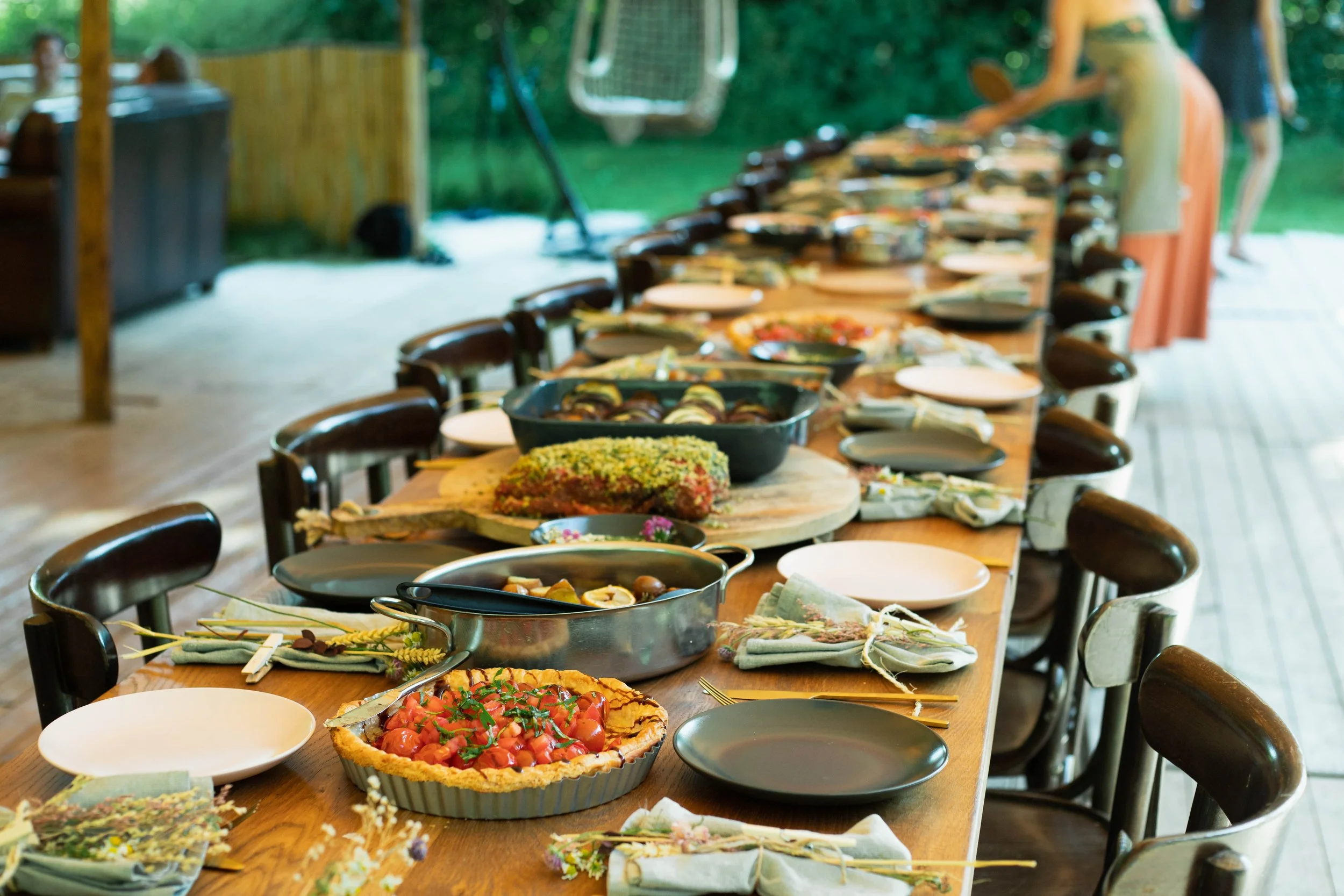 Long wooden dining table set with various dishes, plates, and napkins, outdoors under a canopy or tent, with chairs along both sides, ready for a meal.