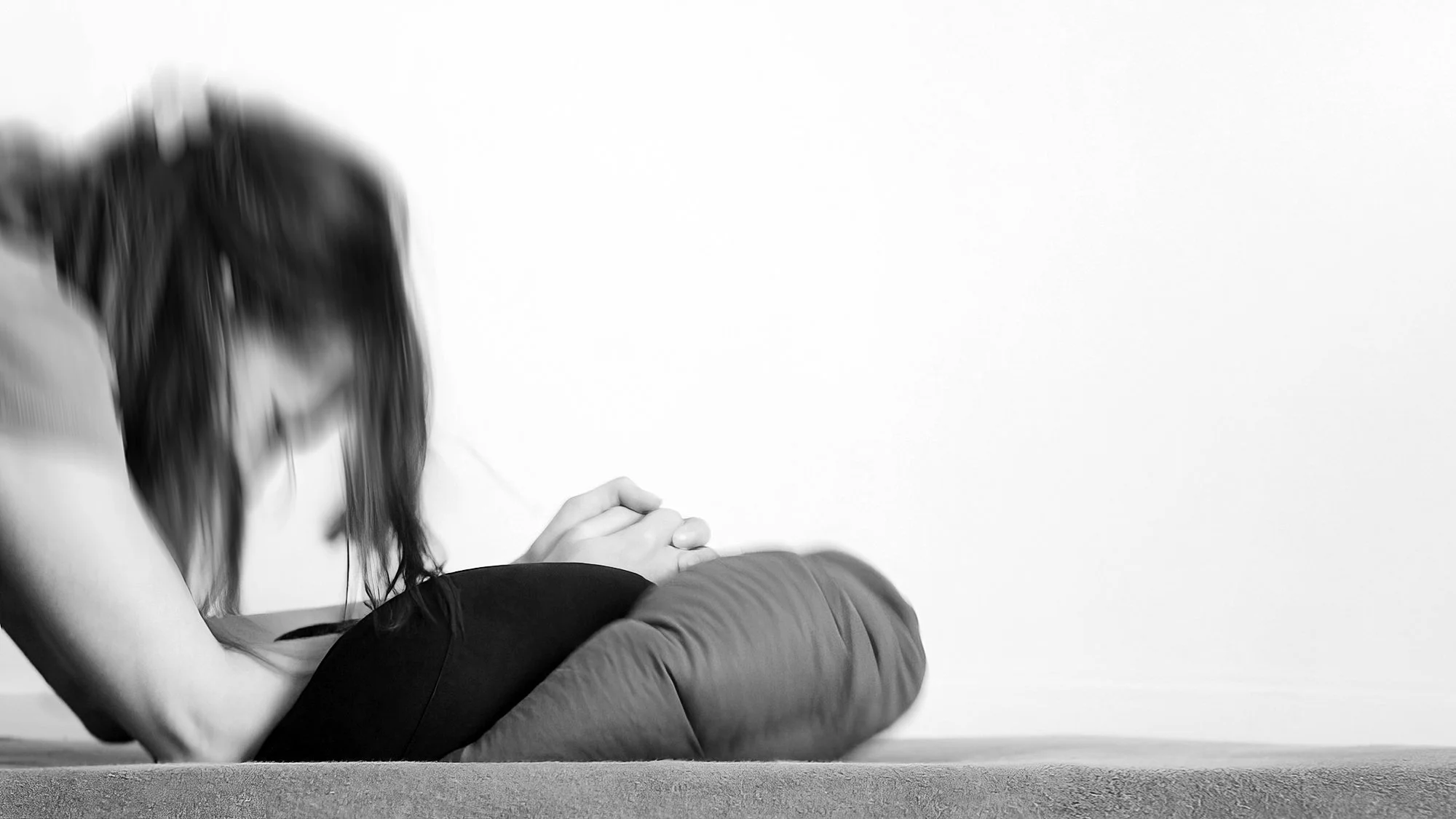 A person is kneeling on a surface with their hands clasped together and resting on their lap, facing downward with their head bowed.