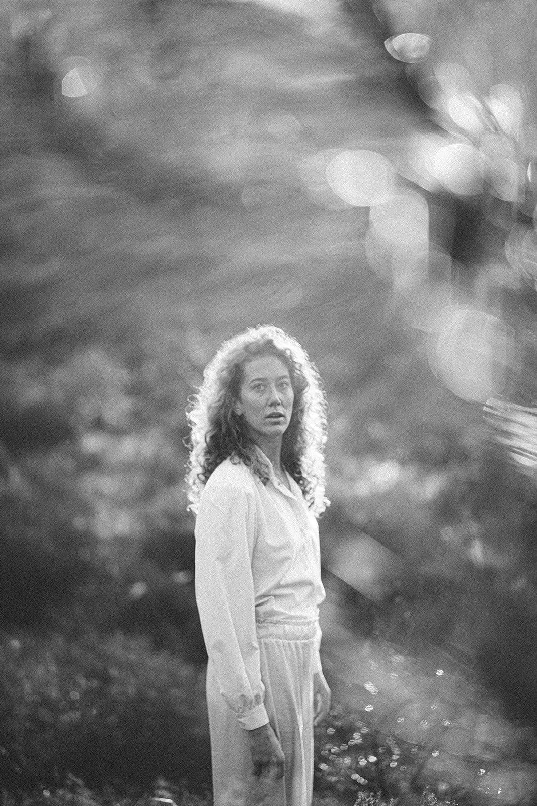 A woman standing outdoors near water, with light reflecting in the background, in black and white.