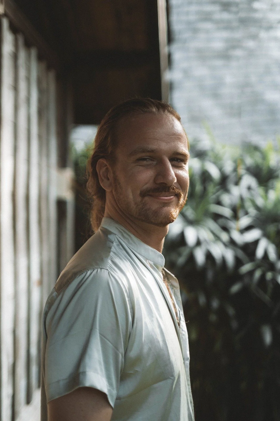 A man with long hair and a beard smiling, wearing a shiny light-colored shirt, standing outdoors near a wooden wall and green foliage, with a background of a gray building.