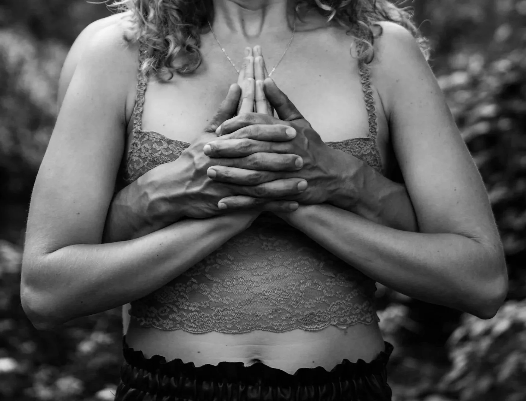 A woman with curly hair wearing a lace top and a necklace stands with her hands pressed together in a prayer or meditation pose, outdoors, in black and white.