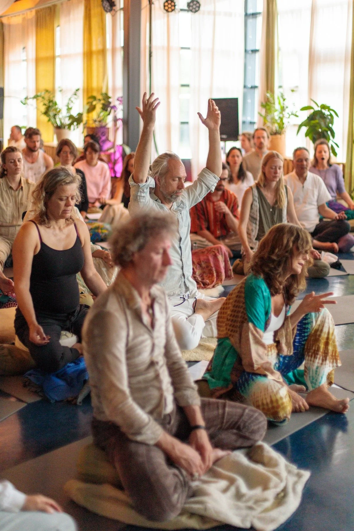 People participating in a yoga or meditation class, with one man raising his arms, sitting on mats in a bright room.