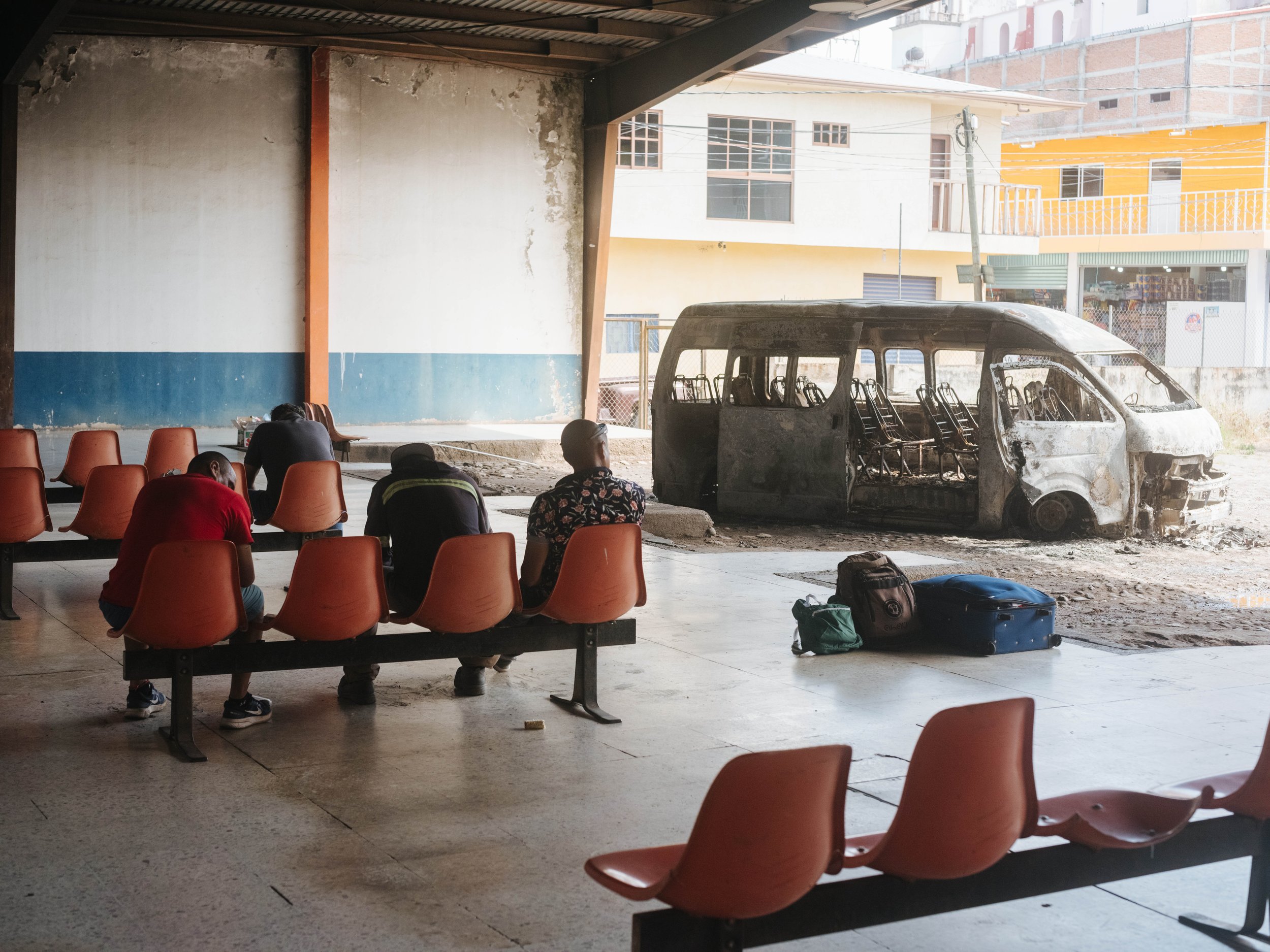 People wait inside a bus terminal in Aguililla, Michoacán, next to a burned vehicle, where daily life continues alongside the remnants of violence.