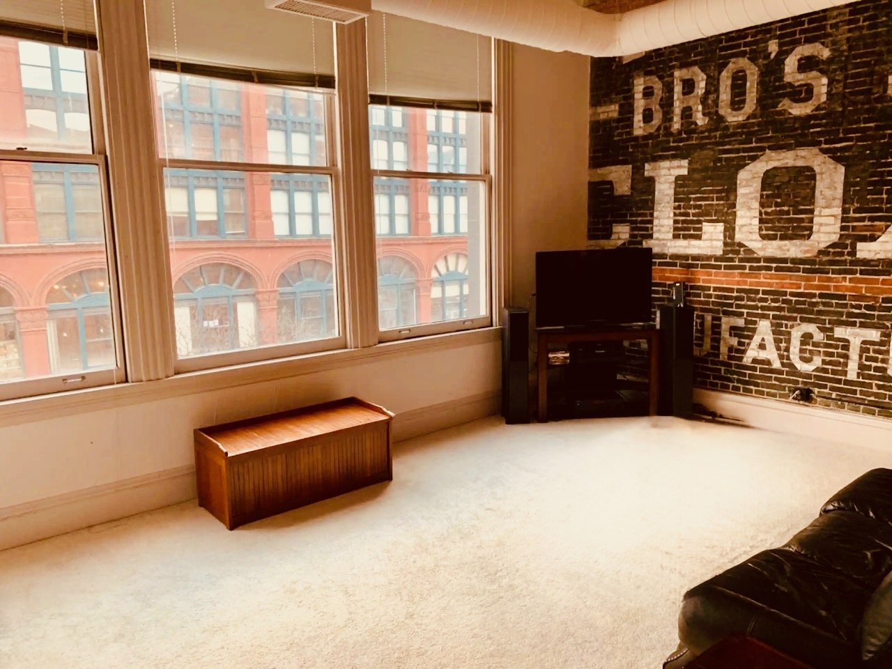 A room with large windows showing red brick buildings outside. Inside, there is a black television on a wooden stand, a black leather sofa, and a wall with a large painted brick mural that reads "BRO'S TO FACT."