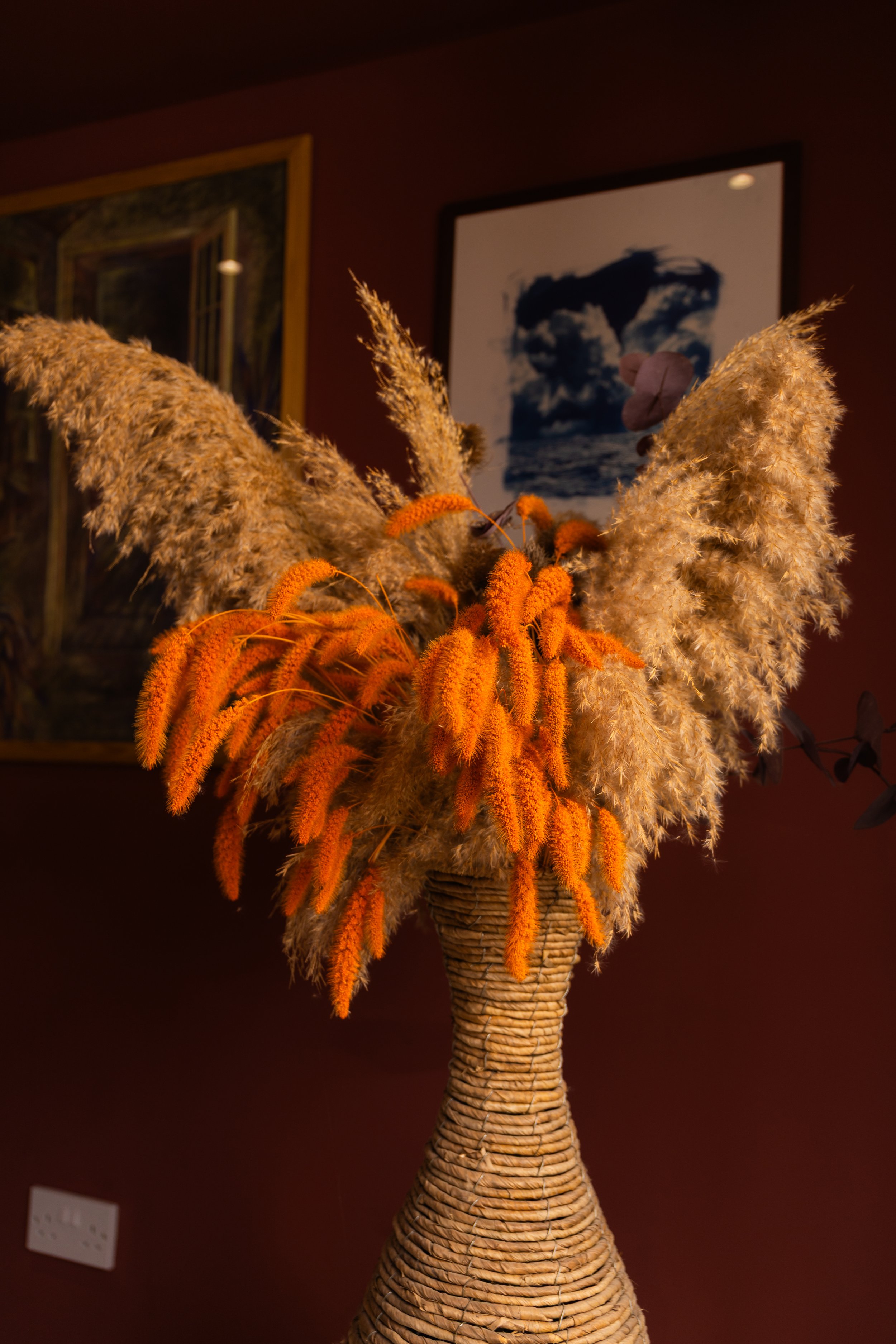 A vase with dried grasses and orange flowers against a dark background.
