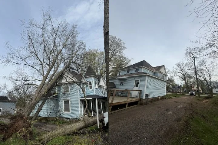 A house with a large fallen tree leaning against it, causing visible damage, on the left side of the image. The house is light blue with multiple stories and a turret. On the right side, the same house appears undamaged from a different angle, showing the same light blue exterior and complex roof structure.