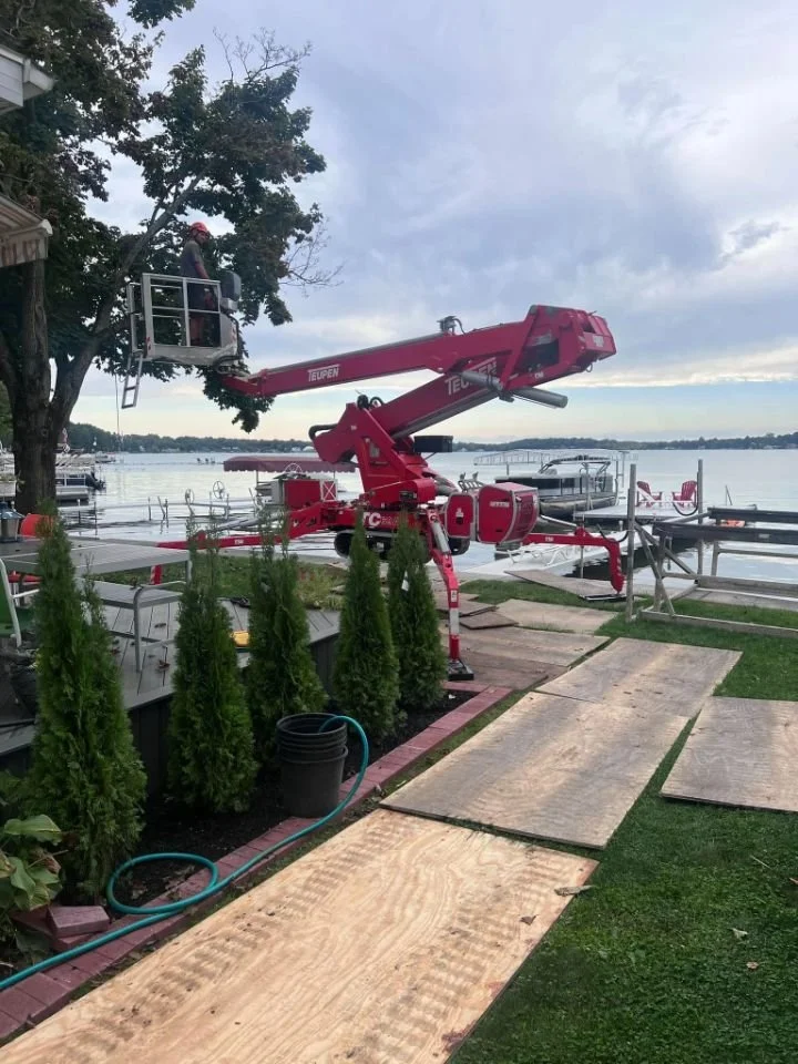 A person in safety gear is using a cherry picker lift to prune a tree near a waterfront. There is a boat dock and water in the background, with multiple boats and a cloudy sky.