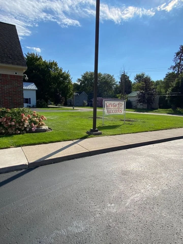 A small yard with green grass, pink flowers, a sidewalk, and a sign that reads 'Let's Go Falcons' with an arrow pointing to the right, under a partly cloudy blue sky.