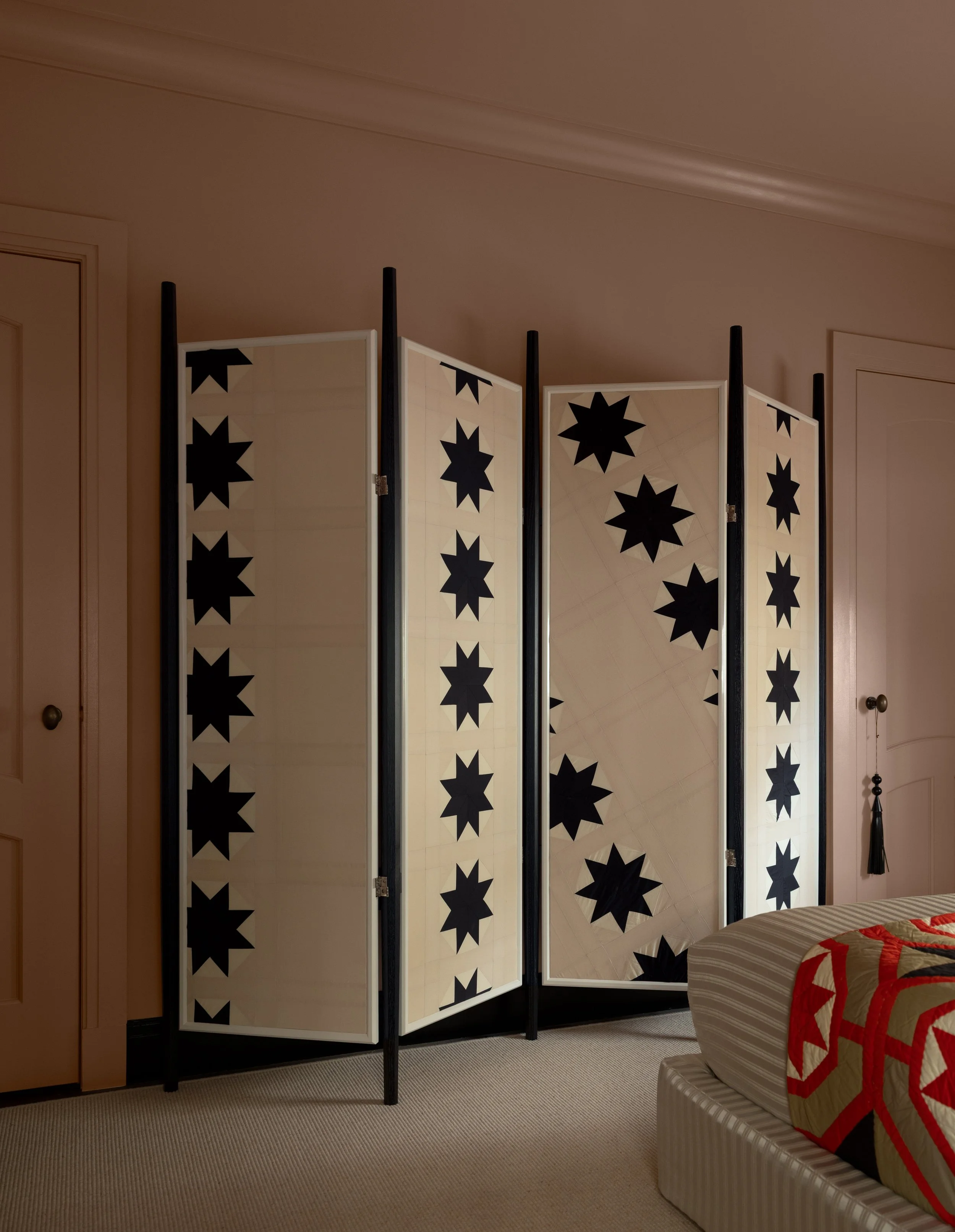 A room divider with black star patterns on cream-colored panels, standing on a beige carpet in a room with pink walls and a closed door. Part of a bed with a quilted bedspread is visible in the corner.