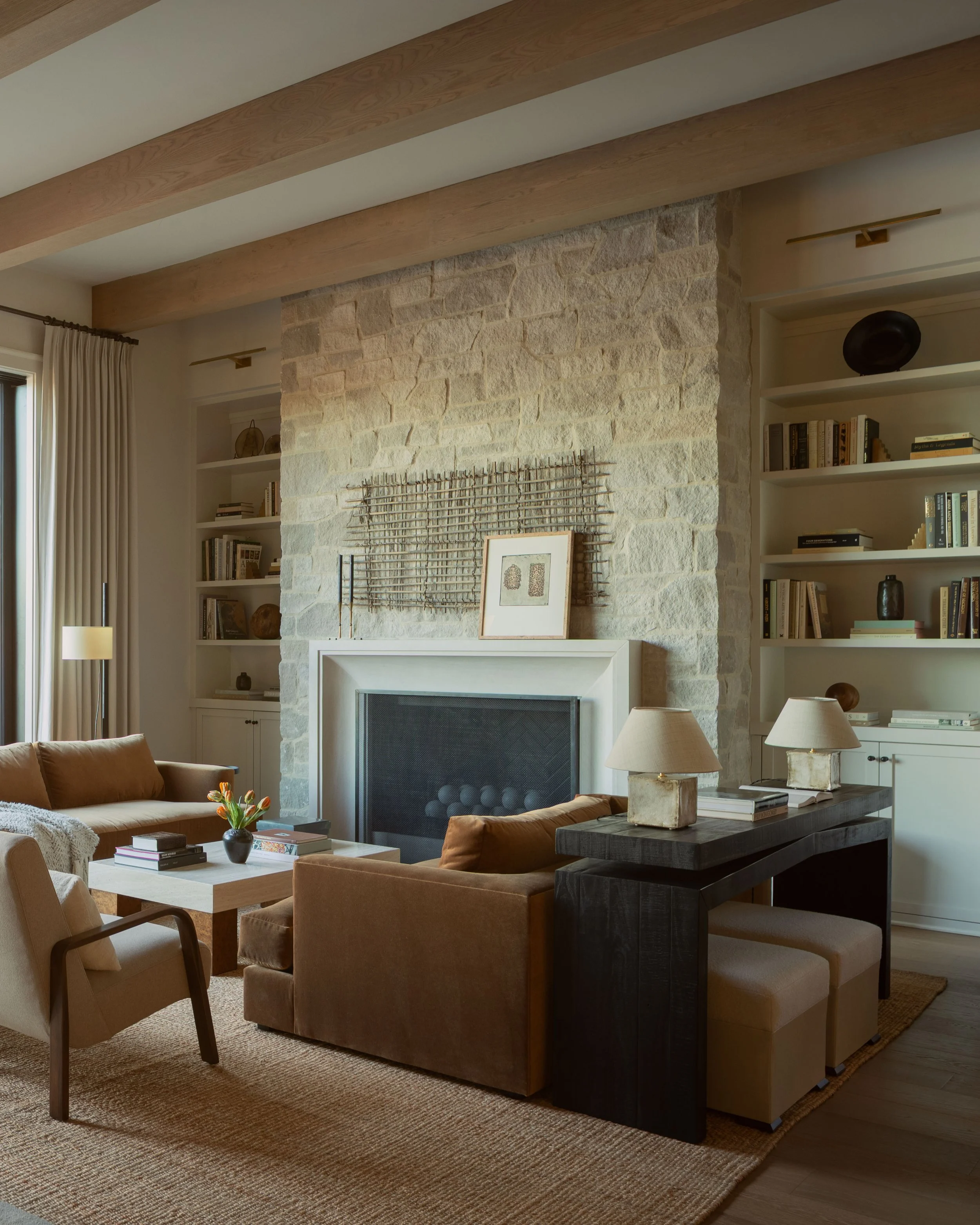 Living room with a stone fireplace, built-in bookshelves, beige sofa, armchair, coffee table with books and a flower vase, wooden ceiling beams, and light-colored curtains.