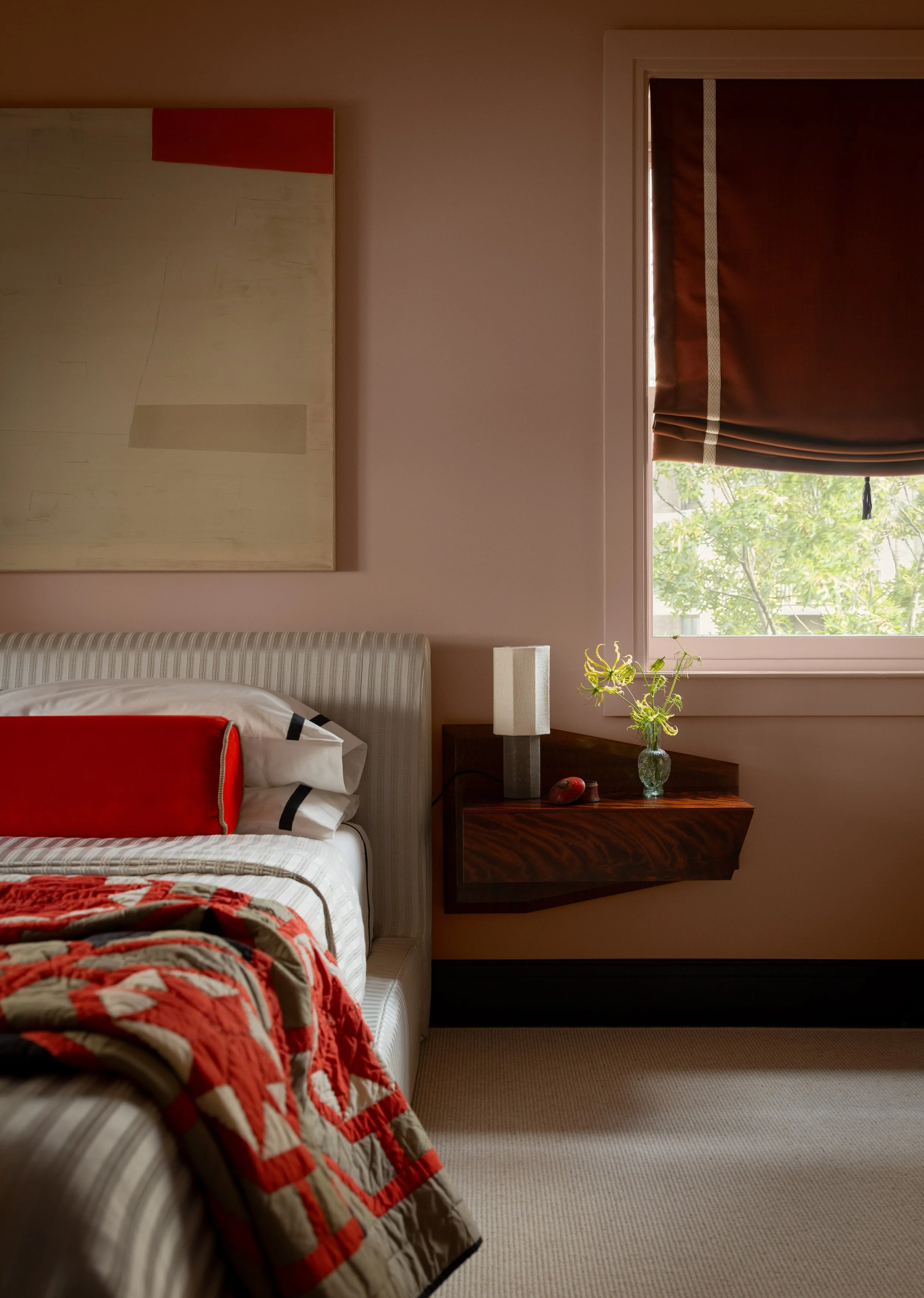 A bedroom with a bed, nightstand, window with brown curtain, and a wall hanging. The bed has a white and beige striped headboard, white sheets, and a red and beige quilt. A pillow with a white pillowcase and a smaller decorative cushion with red, black, and white geometric patterns are on the bed. The nightstand has a vase with yellow flowers, a gray lamp, and a red object. Outside the window, tree branches with green leaves are visible.