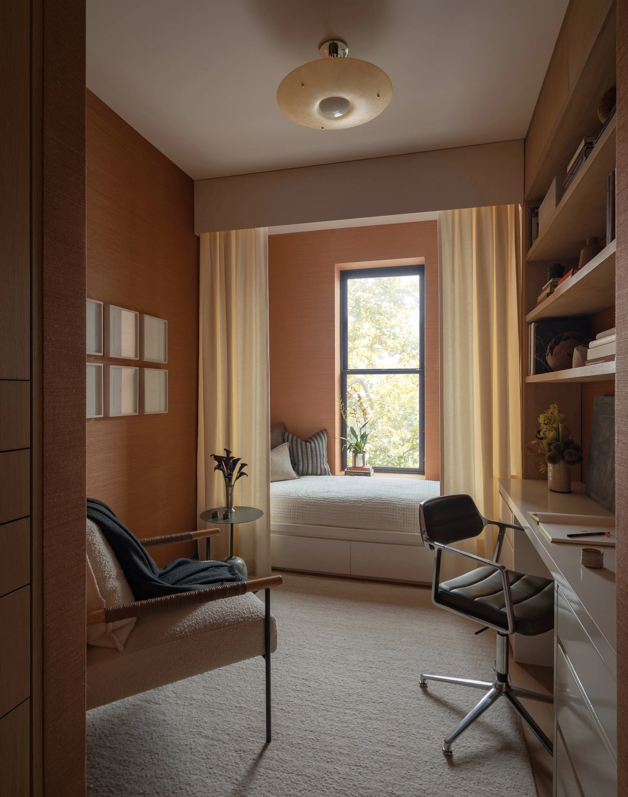 A cozy corner room with a large window, beige curtains, and built-in shelves filled with books and decor items. There's a small bed with cushions, a side table with a potted plant, a gray lounge chair, and a white desk with a black chair. The ceiling has a modern circular light fixture, and the walls are finished in warm wood tones