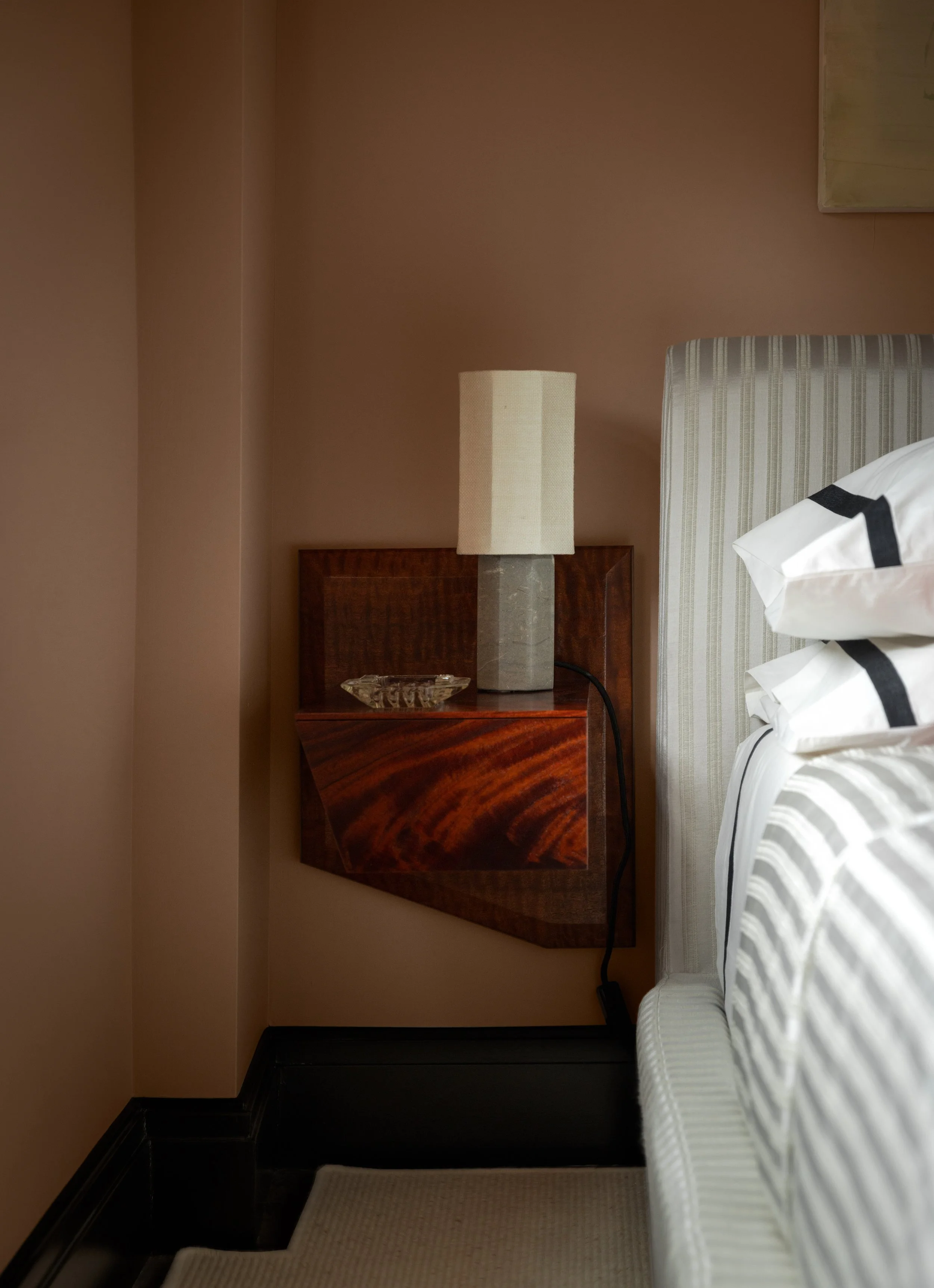 A hotel room with a beige wall, a wooden headboard, a cream-colored lampshade on a concrete base, a fish-shaped glass dish, a bed with white and gray striped bedding, a beige carpet, and a painting on the wall.