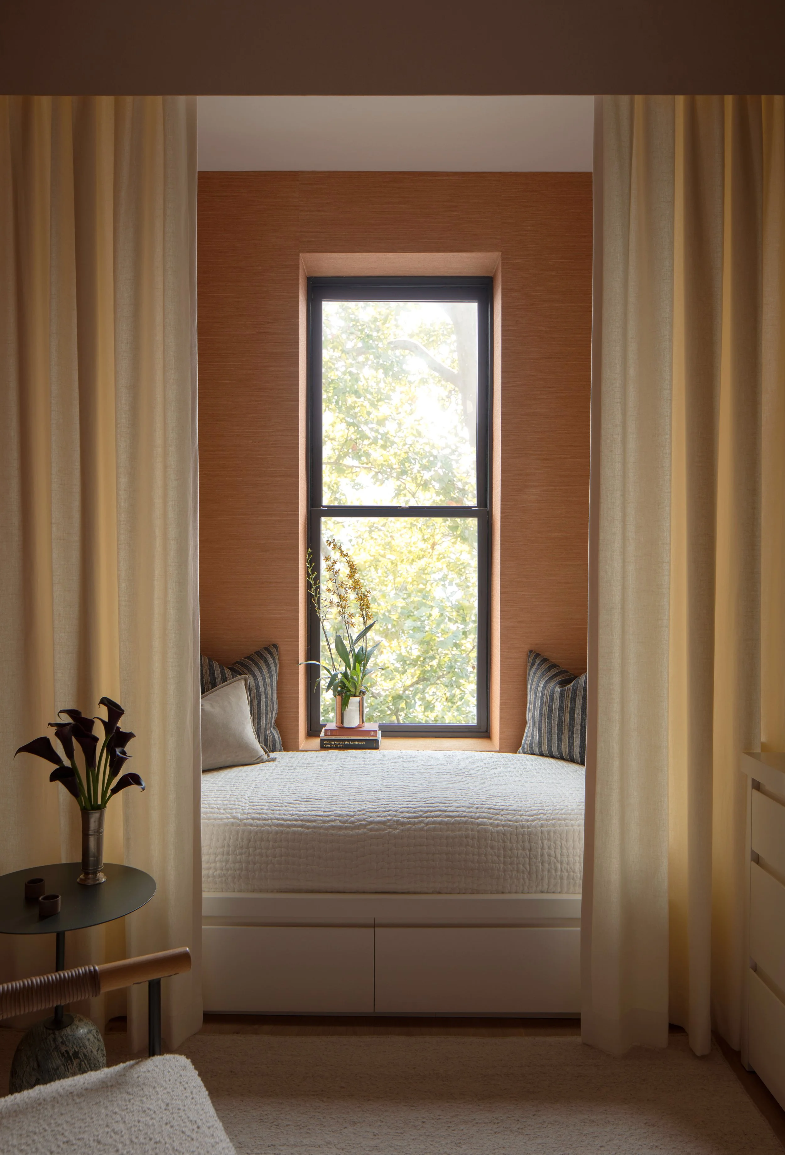 A cozy bedroom with a built-in window seat surrounded by beige curtains, a large window with views of green trees, a small flower arrangement on a side table, and striped throw pillows on the window seat.