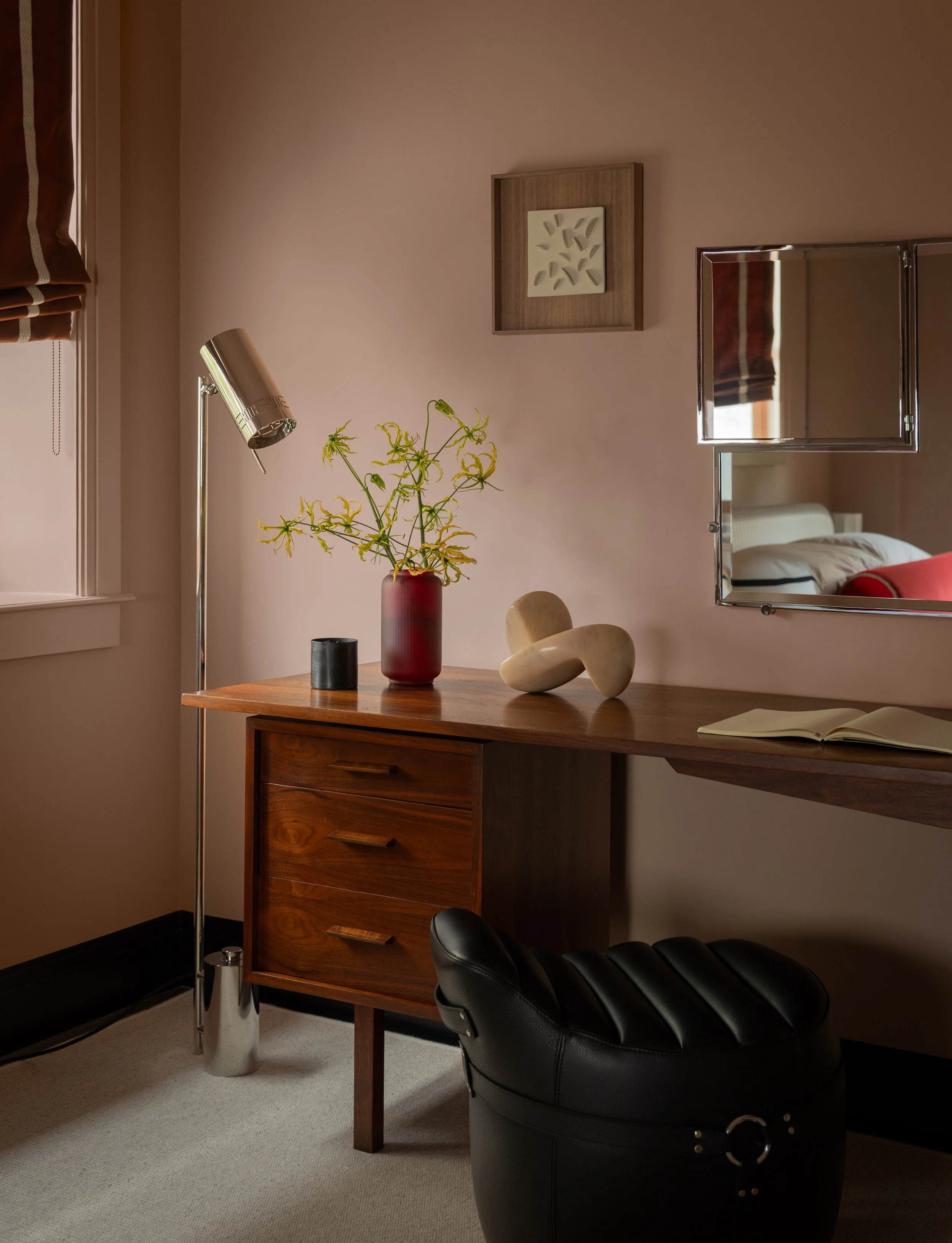Living room with a wooden desk, a black leather chair, a vase with yellow-green flowers, a wall mirror, framed artwork, a floor lamp, and a television partially reflected in the mirror.