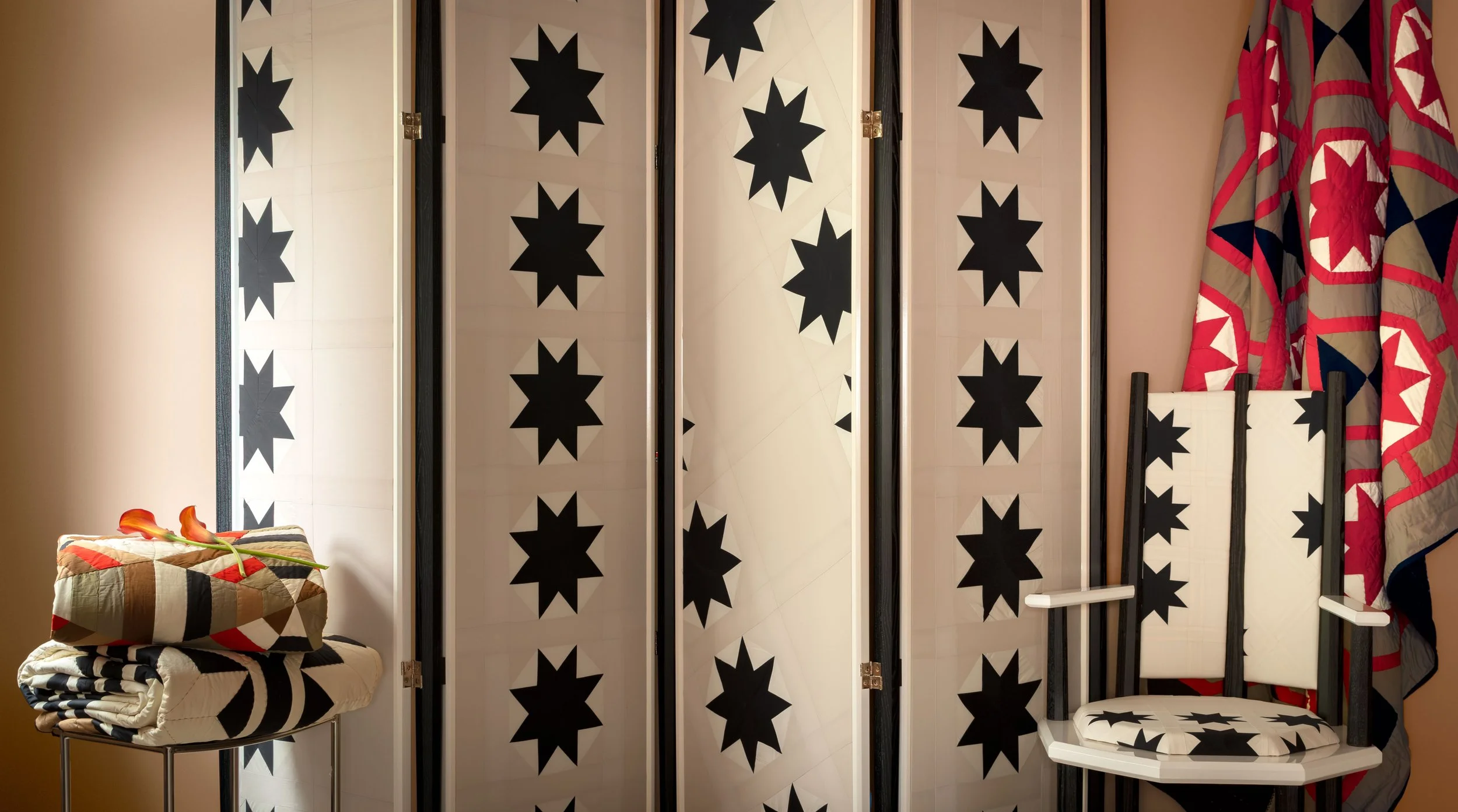 A folding room divider with black starburst patterns on white panels, positioned between a beige wall and red, blue, and beige patterned curtains. A small striped quilted chair with a pillow on top, and a smaller matching quilted stool with folded quilts are to the left. A white wooden chair with armrests and a striped cushion is on the right.
