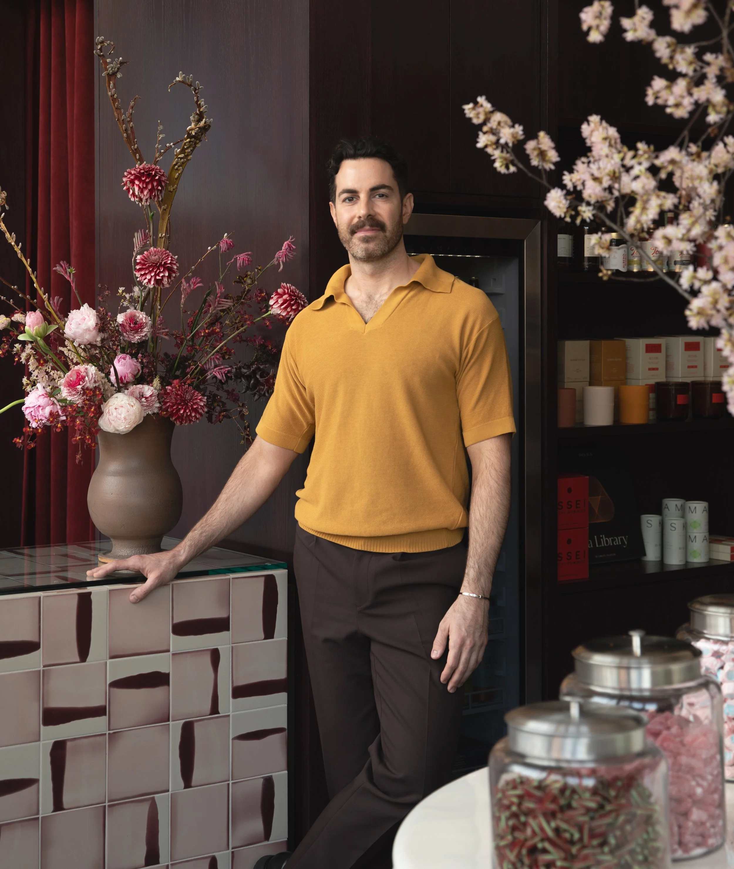 A man with dark hair and a beard wearing a mustard yellow polo shirt and dark pants stands next to a large floral arrangement in a tall beige vase. The arrangement includes pink and red flowers with branches and sprigs. Behind him is a dark wooden wall with red curtains on the left and a black shelf with boxes and jars on the right. There are bowls of pink and brown treat sticks in the foreground.