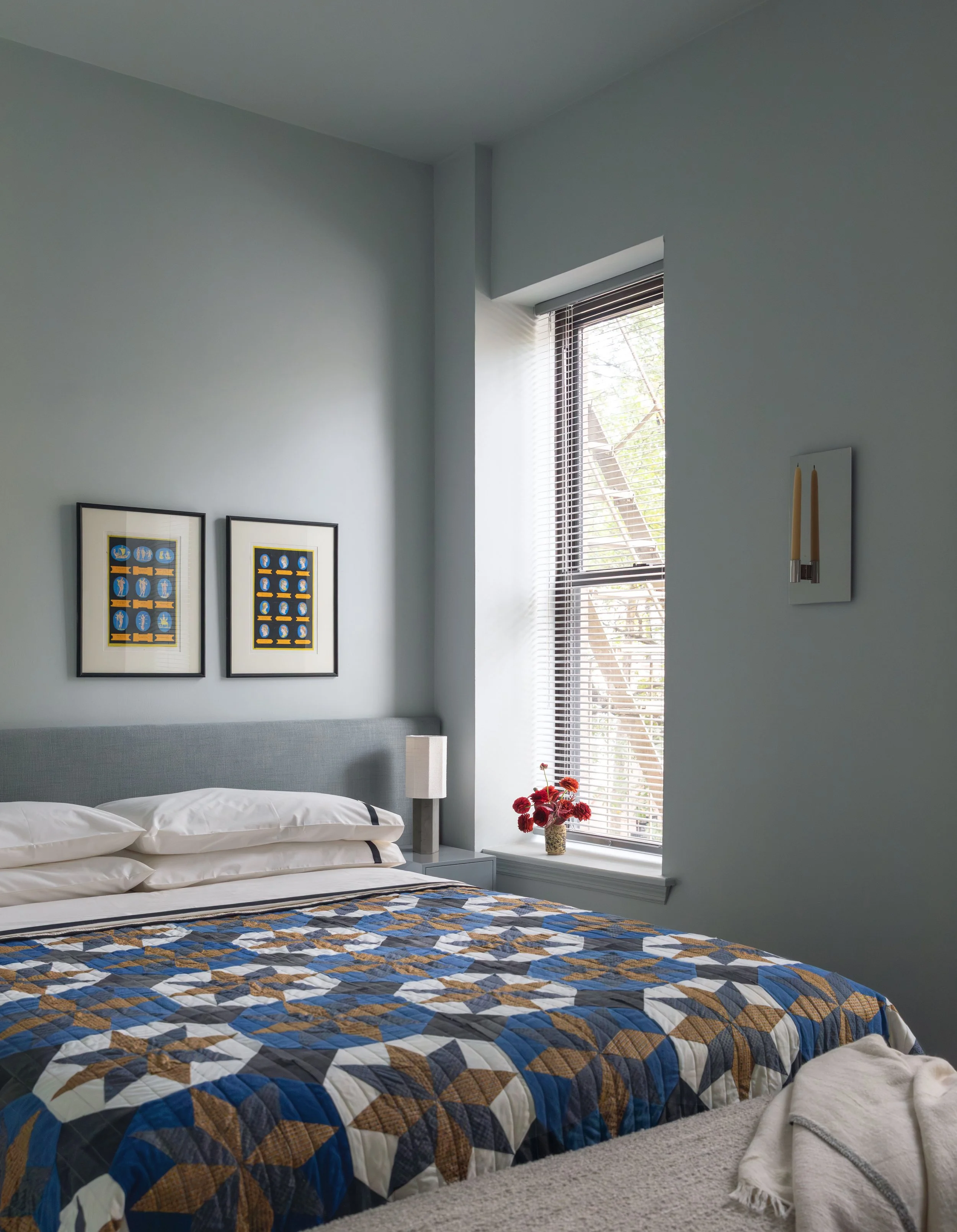 A bedroom with a bed covered in a multicolored quilt, two pillows, a nightstand with a lamp, a vase of red flowers, framed artwork on a light blue wall, and a window with blinds letting in natural light.