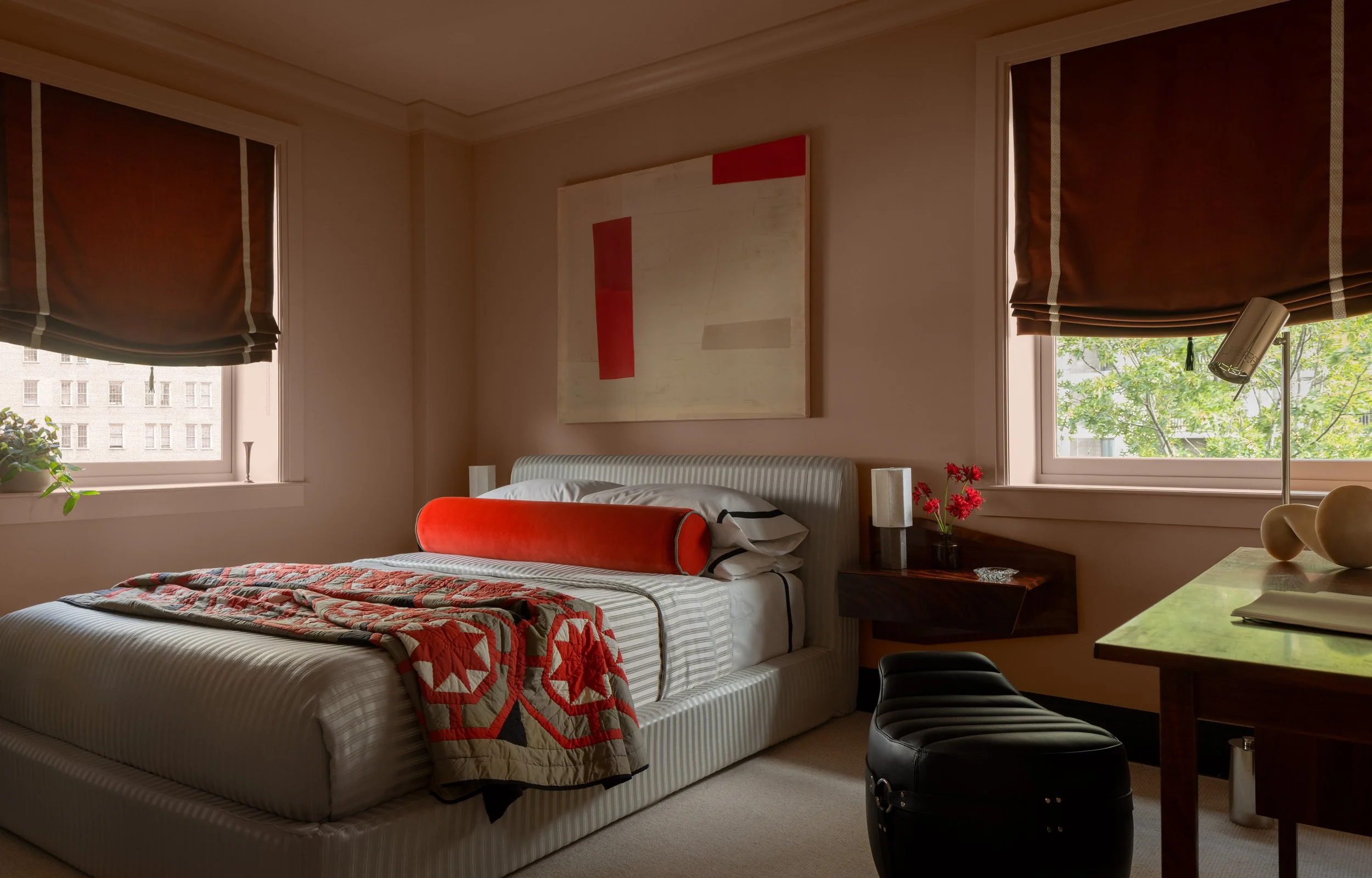 A bedroom with a bed, side table, and two windows with brown curtains. There is artwork on the wall and a table with decorative objects, a lamp, and a chair nearby.