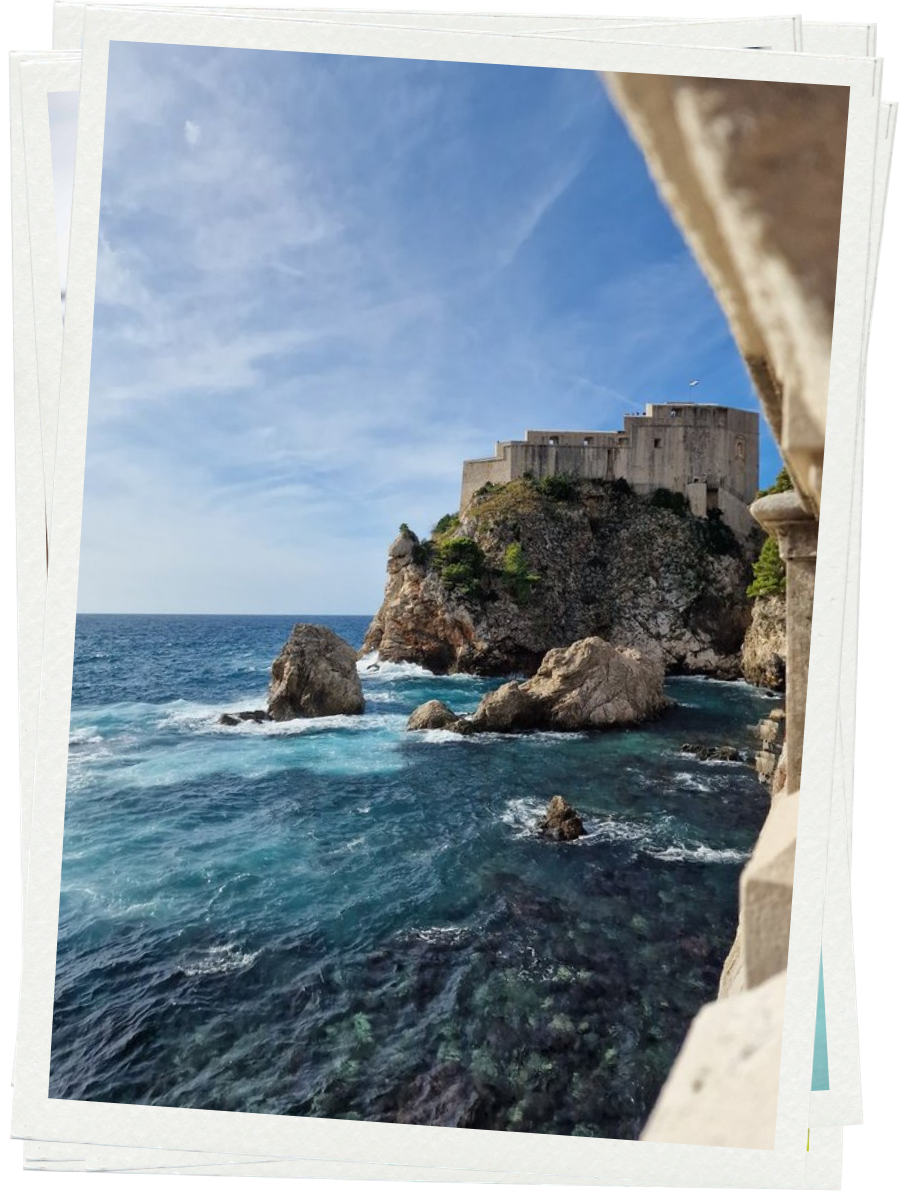 A landscape view of a rocky coastline with a fortress on a cliff, blue ocean waves, and a cloudy sky.