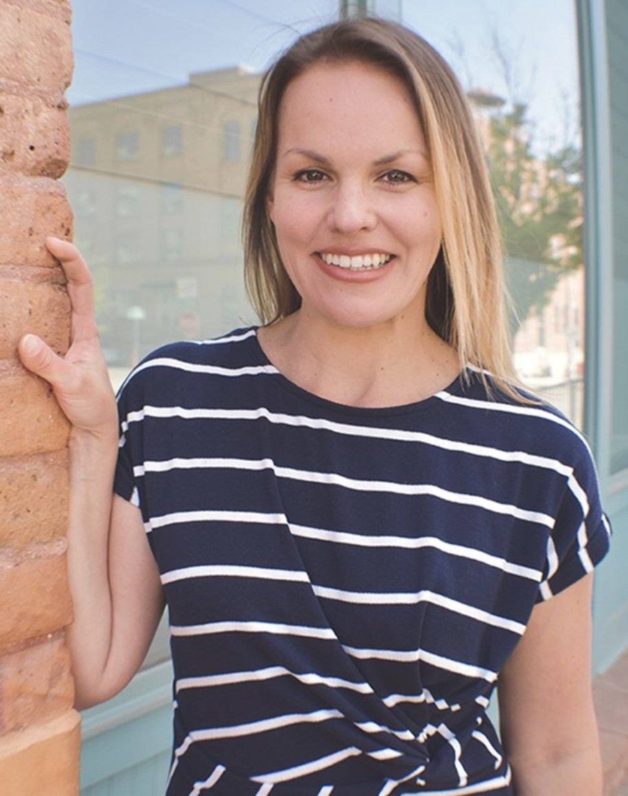 A woman with shoulder-length blonde hair, wearing a navy blue t-shirt with white horizontal stripes, smiling and leaning against a brick wall outside near a glass window.