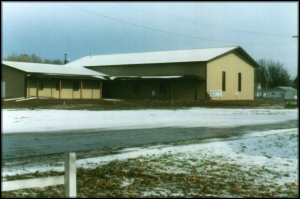 A single-story building with a sloped roof and beige walls, situated alongside a snowy road, with a cloudy sky overhead.