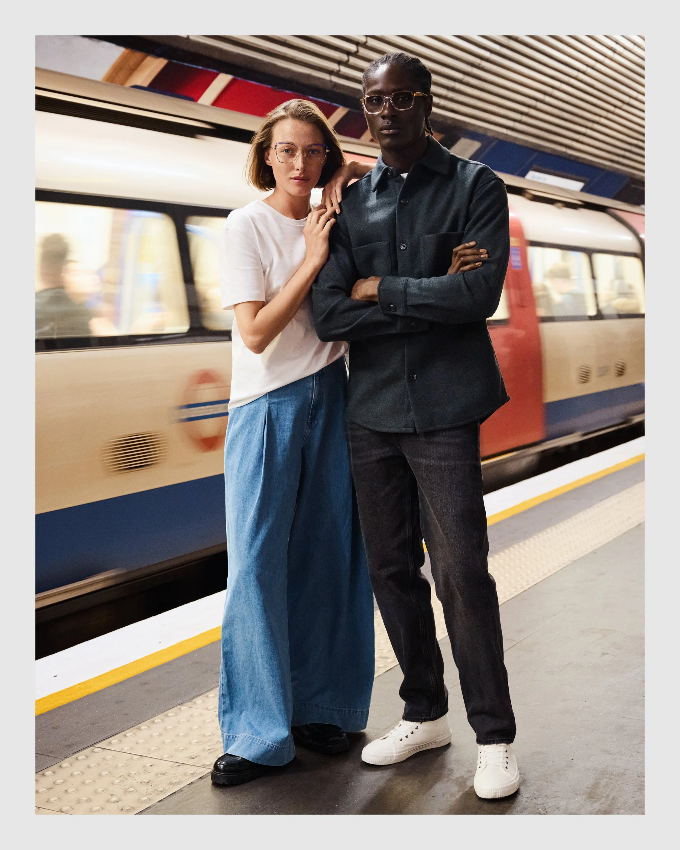 Female and male models wearing William Morris eyewear on a London Underground platform with a moving train behind them.